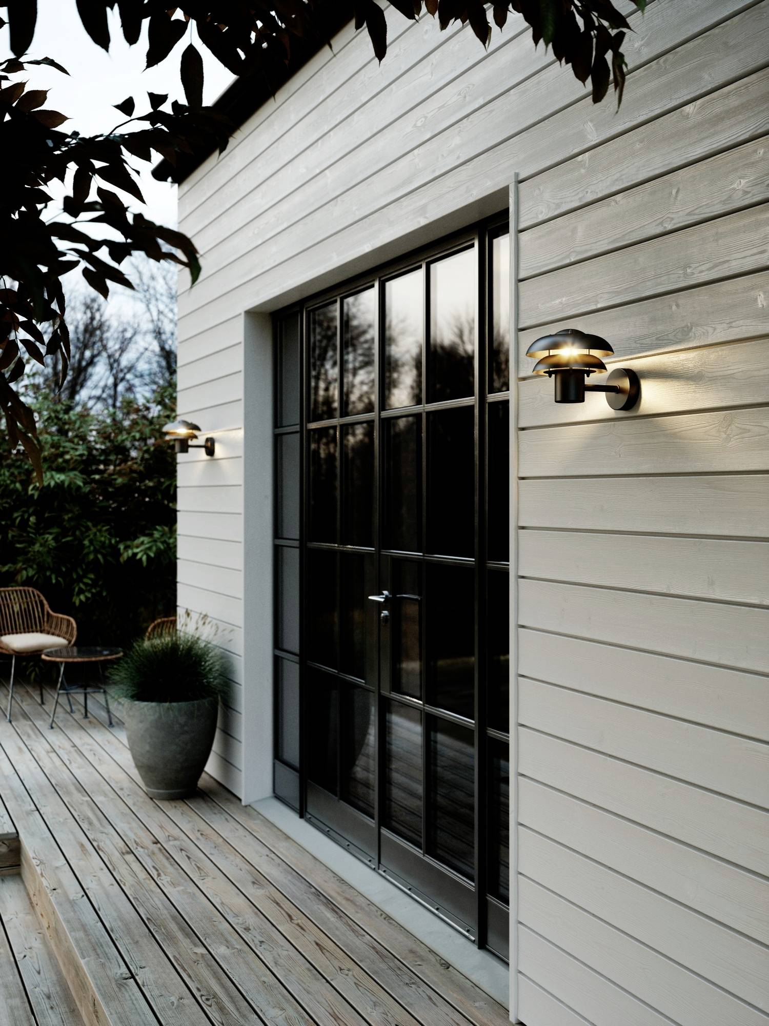 A modern house exterior with large glass doors, wooden siding, and outdoor wall lighting. A wooden deck and potted plant are visible.