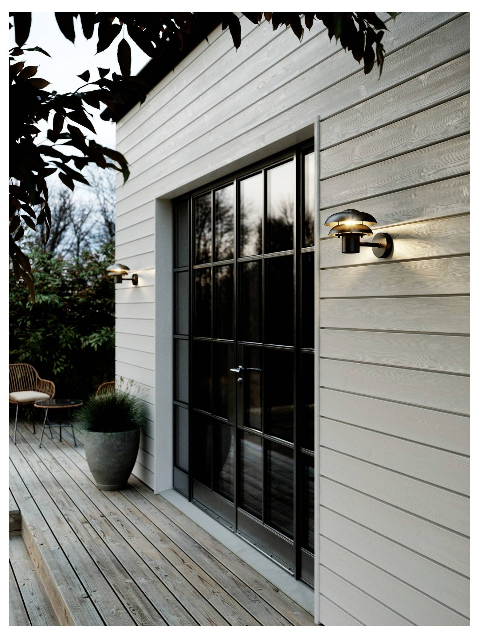 A modern house with wooden siding and a glass door, framed by outdoor wall lights. A small deck and potted plant are visible.