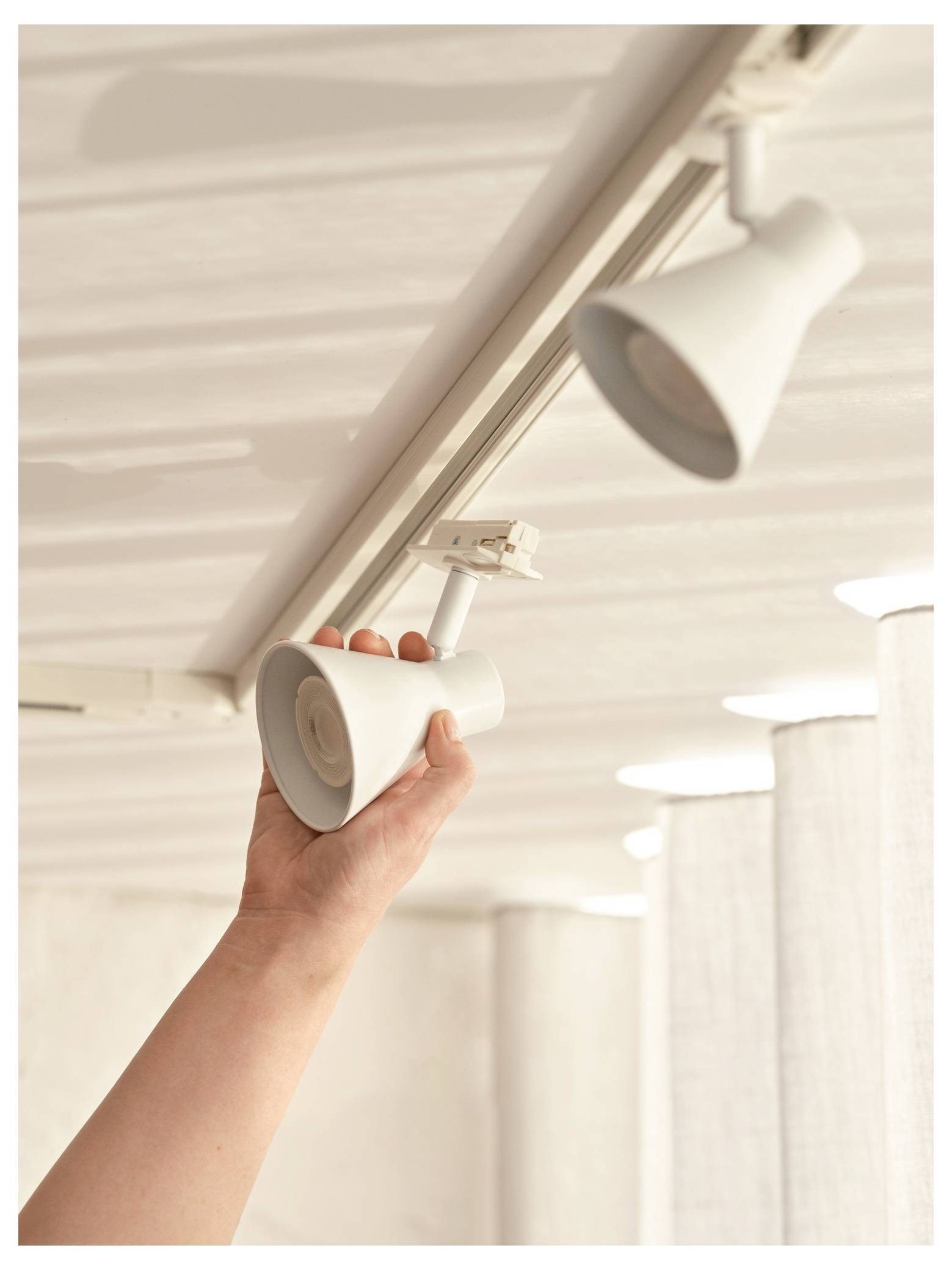A person adjusts a white track light on a ceiling, surrounded by soft curtain light in the background.