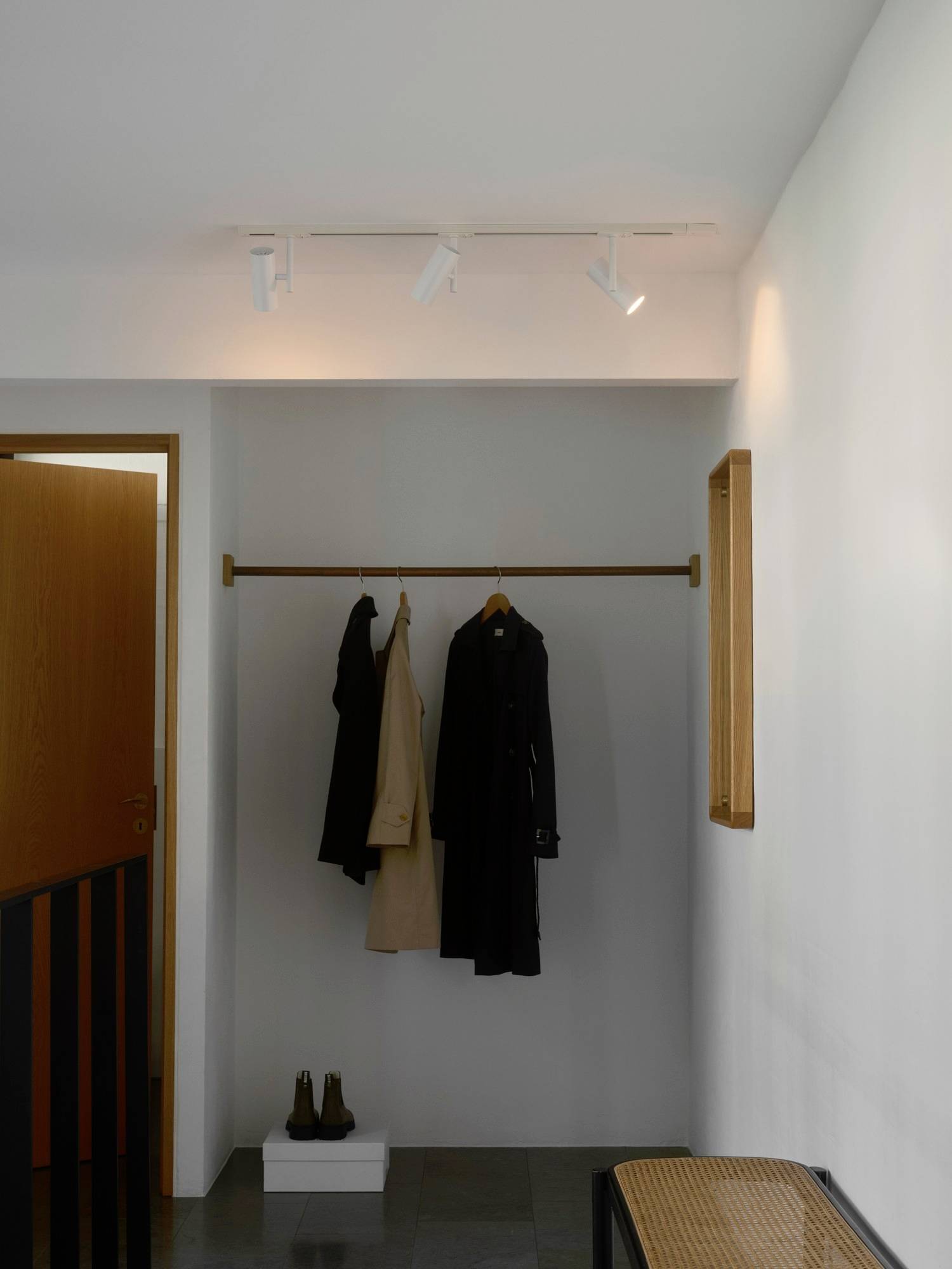 A minimalist hallway with a wooden coat rack holding two coats. Below, a pair of boots sits on the floor, with soft overhead lighting.