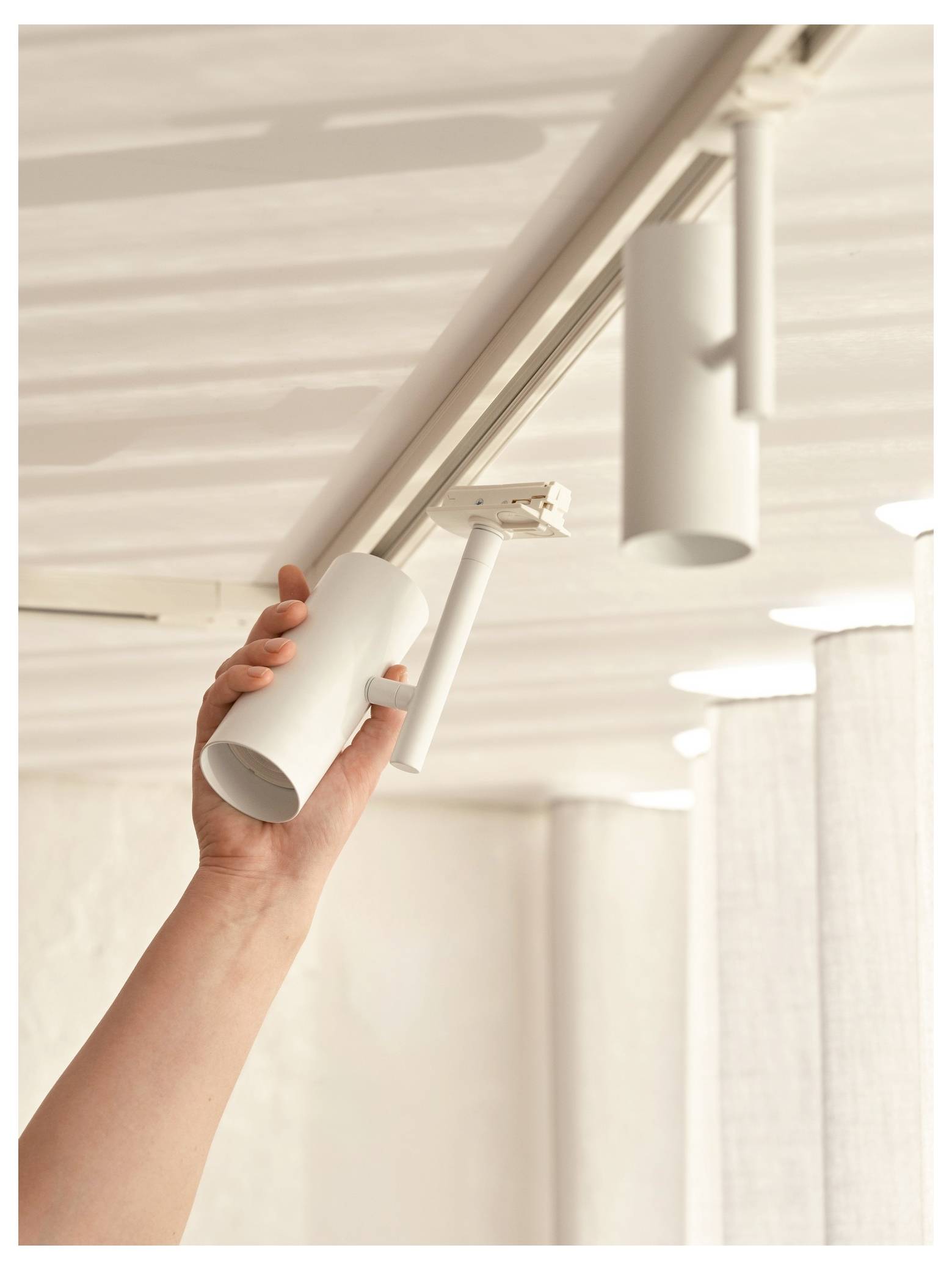 A hand adjusting a white cylinder-shaped track light on a ceiling rail, with sunlight filtering through sheer curtains in the background.
