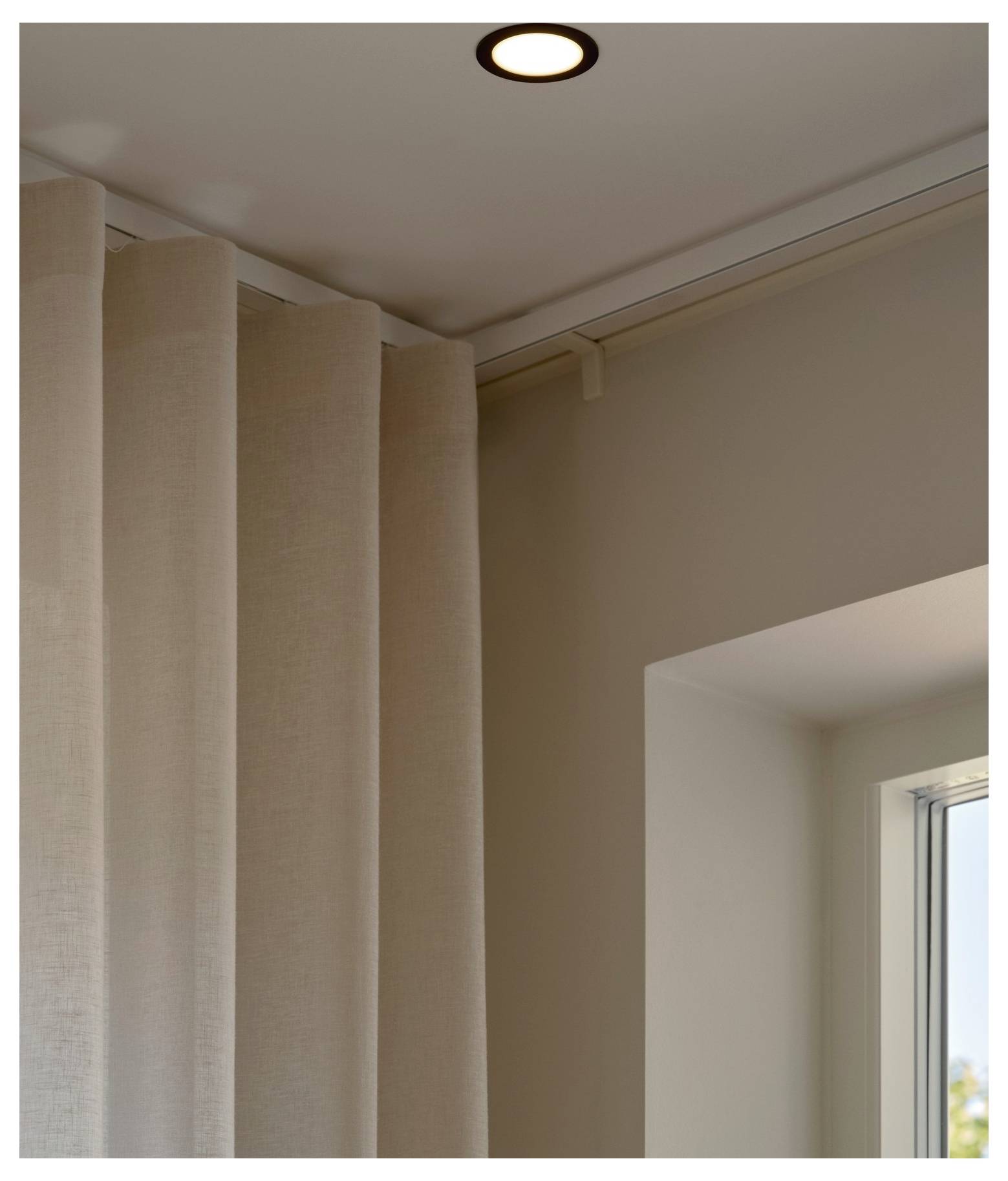 A corner of a room showing beige curtains, a ceiling light, and part of a window with a view of the sky.