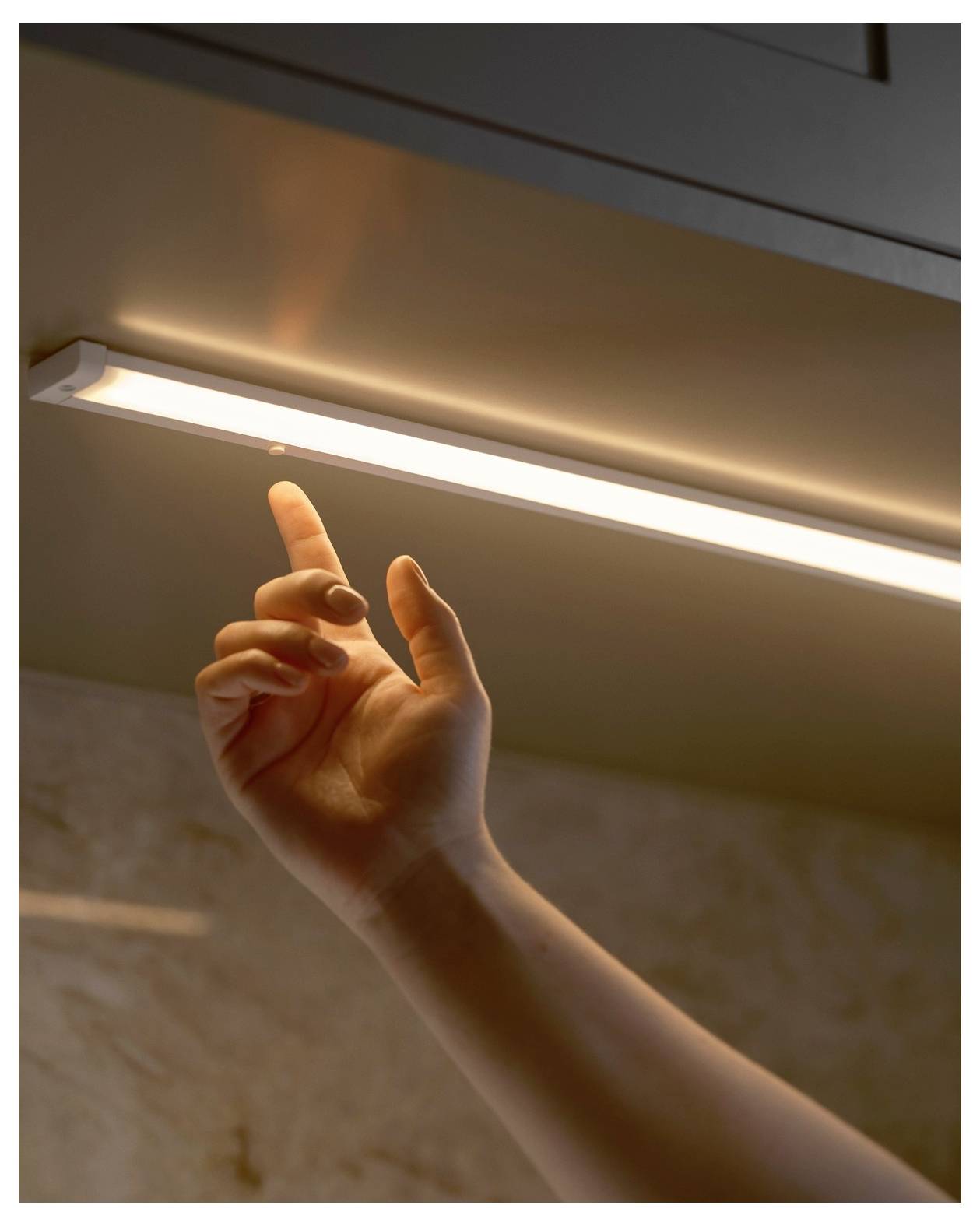 A hand reaches to turn on a strip light under a cabinet, illuminating a kitchen countertop.