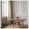 A minimalist room with a wooden table and chair, an open book and pen on the table, pendant light above, and tall windows with beige curtains.