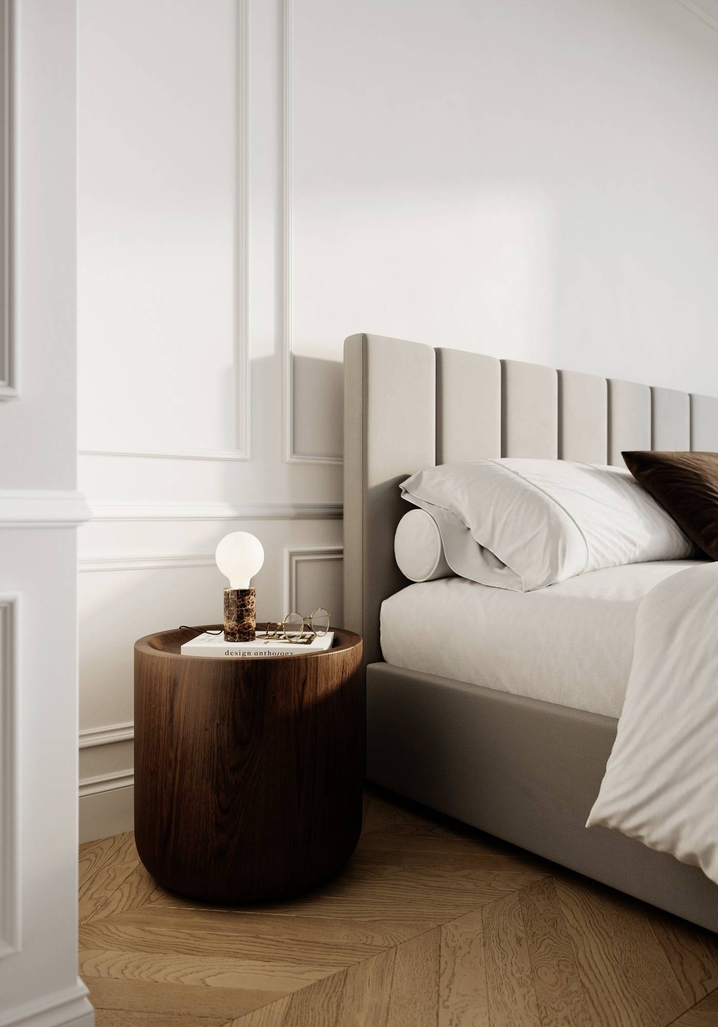 A modern bedroom with a gray upholstered bed and white bedding. A round wooden nightstand holds a light bulb and glassware.