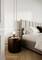 A modern bedroom with a gray upholstered bed and white bedding. A round wooden nightstand holds a light bulb and glassware.