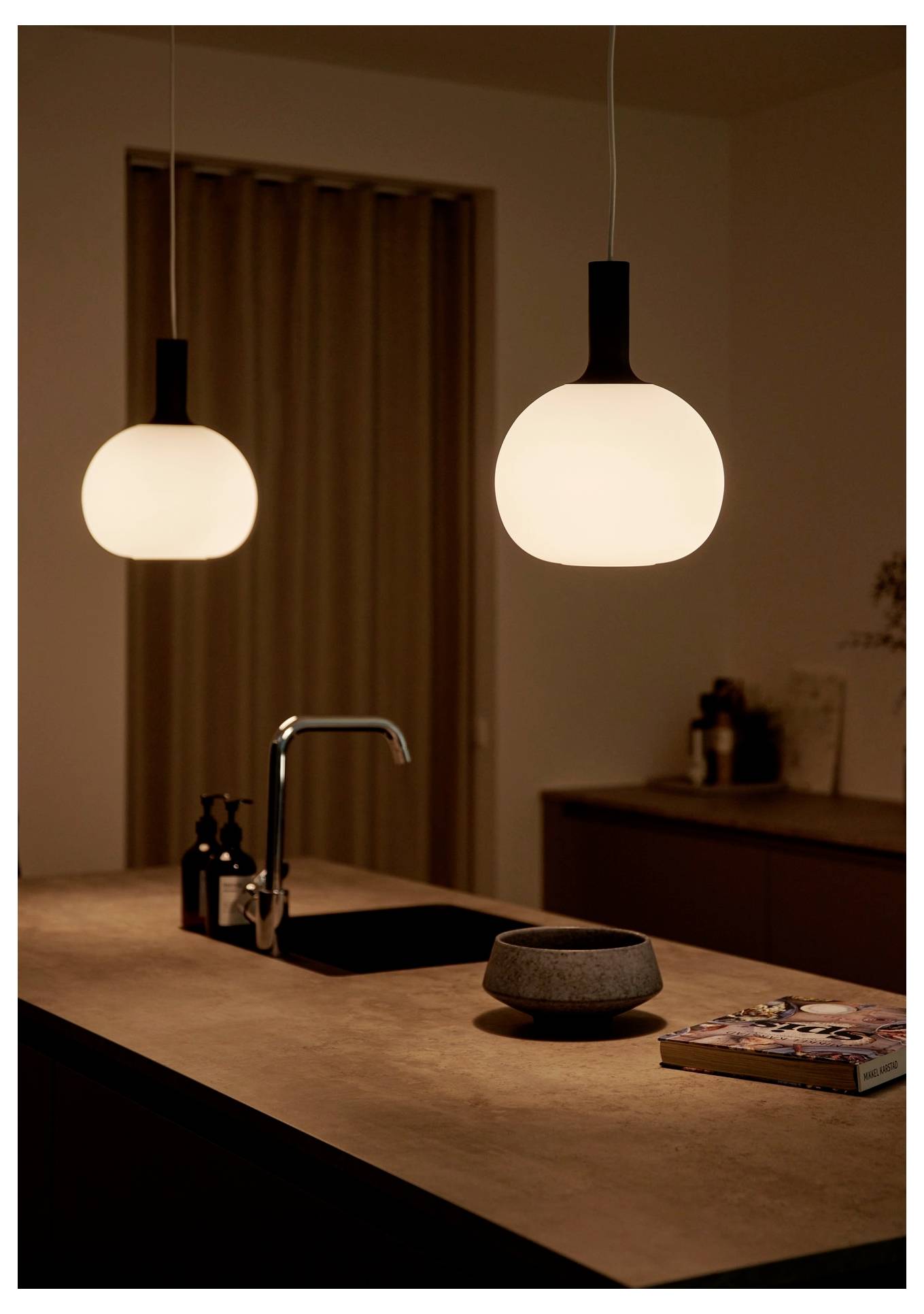A modern kitchen with two glowing pendant lights hangs over a central island with a sink and a cookbook, creating a warm atmosphere.