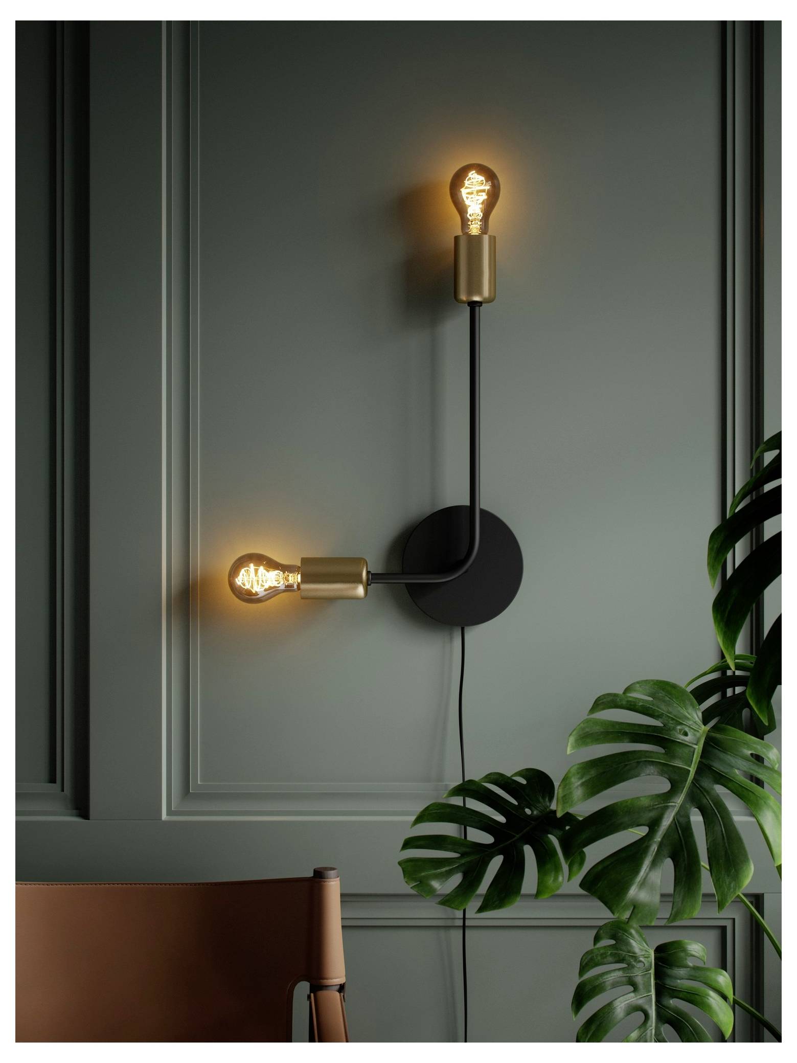 Modern wall sconce with two glowing bulbs on a dark green paneled wall; nearby, a brown chair and a large leafy plant add decor elements.