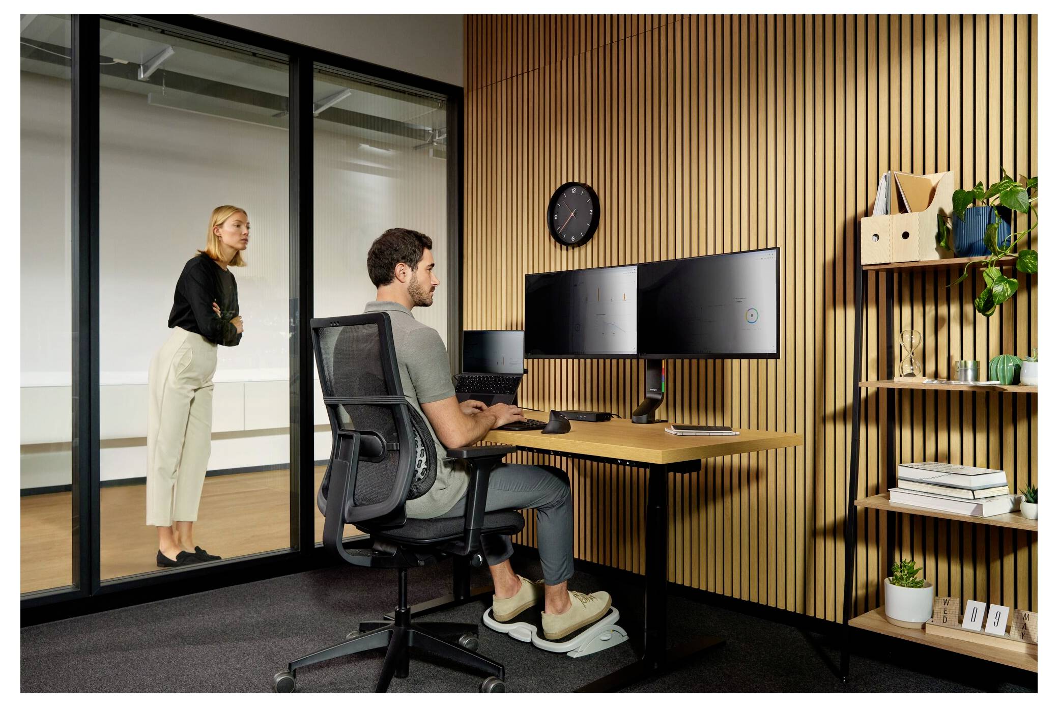 Office scene with man seated at a standing desk using dual monitors while a woman stands in a glass-walled room behind him.