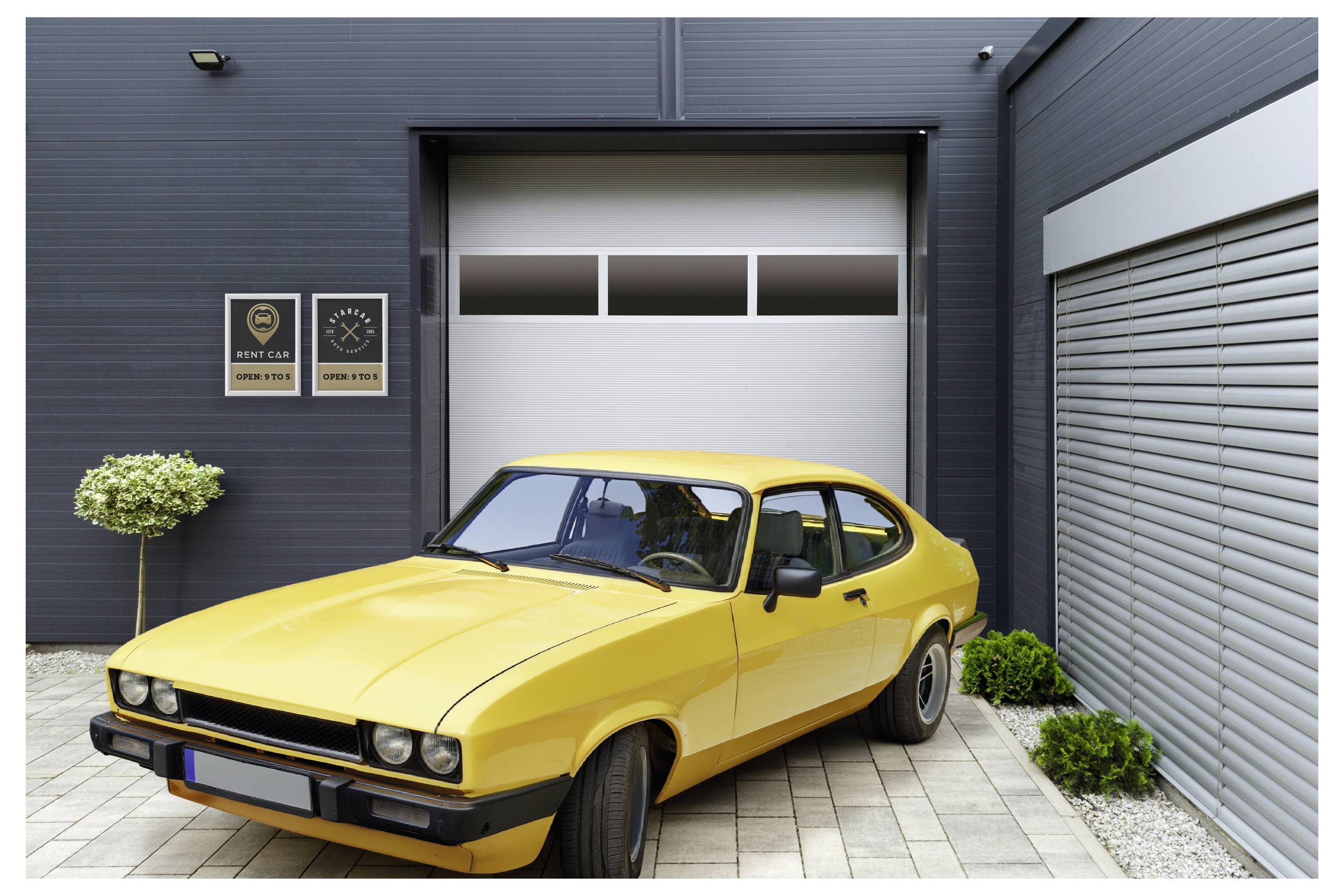 A yellow vintage car is parked on a brick driveway in front of a modern garage with two wall-mounted signs displaying temperature and closed status.