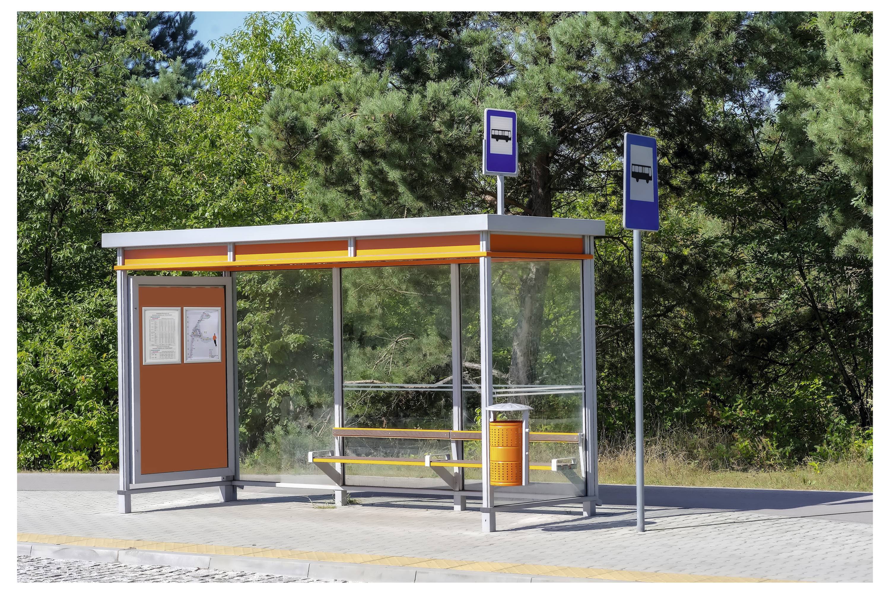 A small bus stop shelter with a bench, map, and signs, situated on a paved area surrounded by trees in the background.