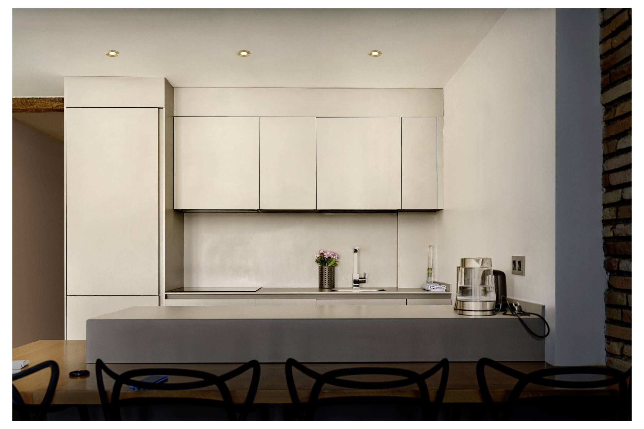 Modern kitchen with light cabinetry, sleek countertops, and a row of black chairs. Appliances and a small plant are neatly arranged.