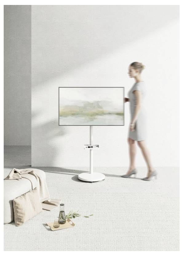A person in a gray dress stands beside a modern TV on a minimalistic stand in a bright, airy room with neutral tones.