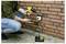 A person is using a yellow cordless drill to fasten a device on a brick wall, highlighting DIY home improvement. A battery and charger are visible on the ground.