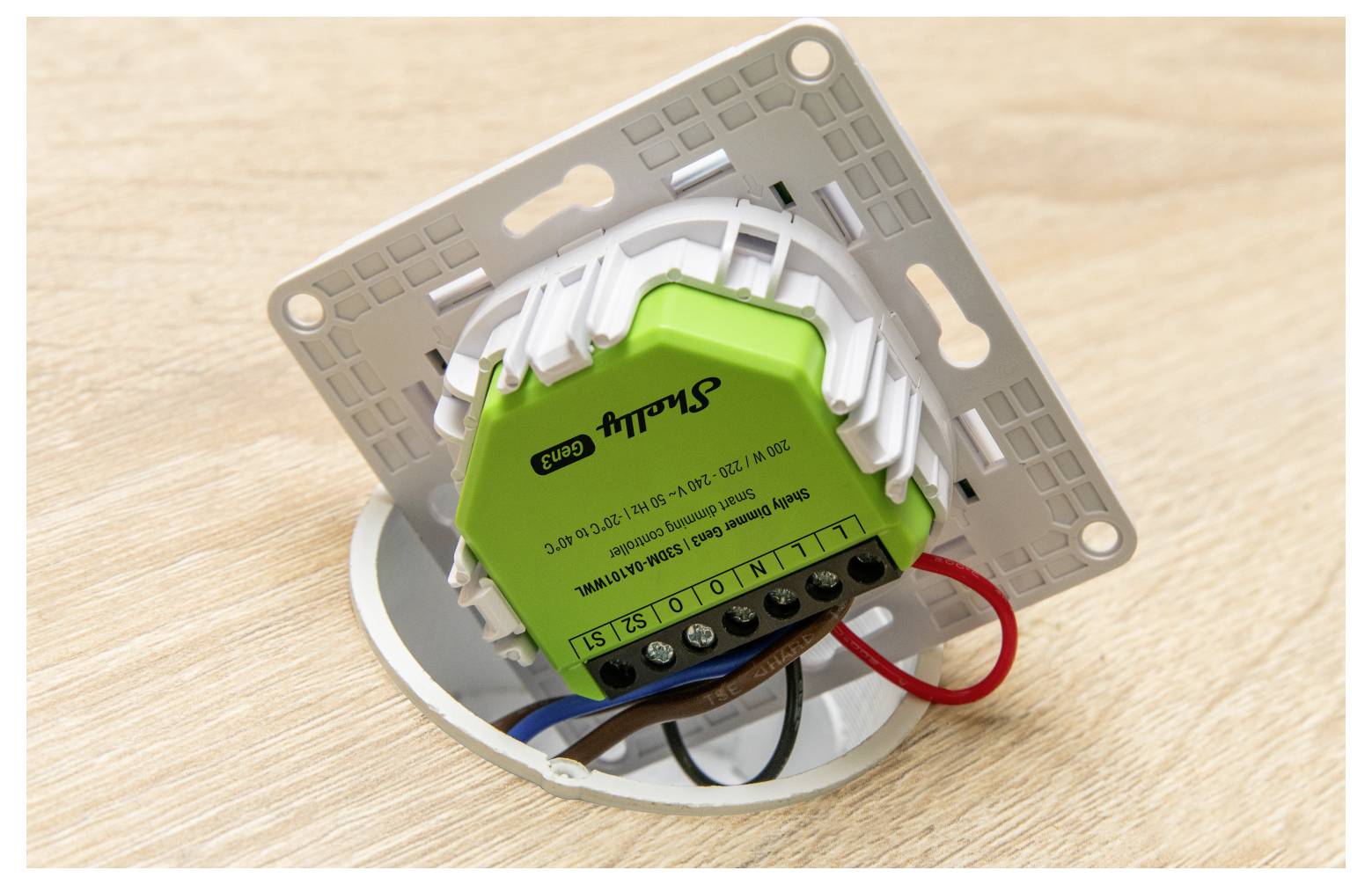 A green smart switch module with wiring connected, mounted on a rectangular wall plate, placed on a wooden surface.