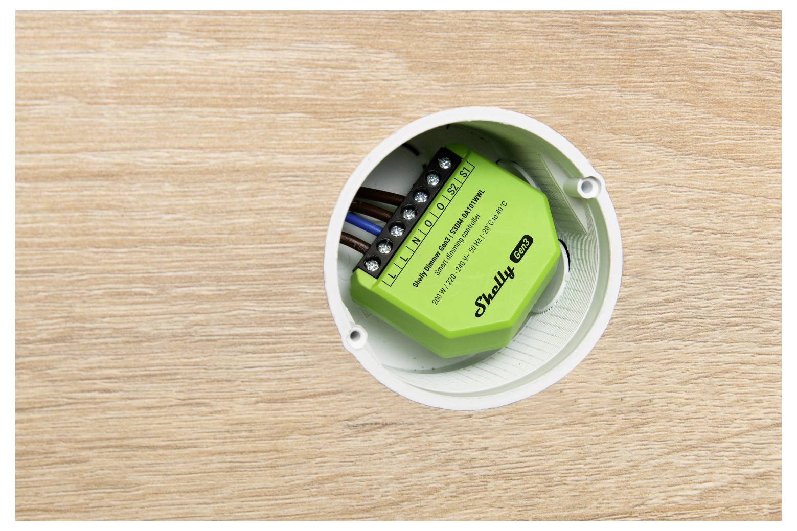 A green Shelly 1PM smart relay is installed inside an electrical box, visible against a wooden surface, showing its wiring connections.