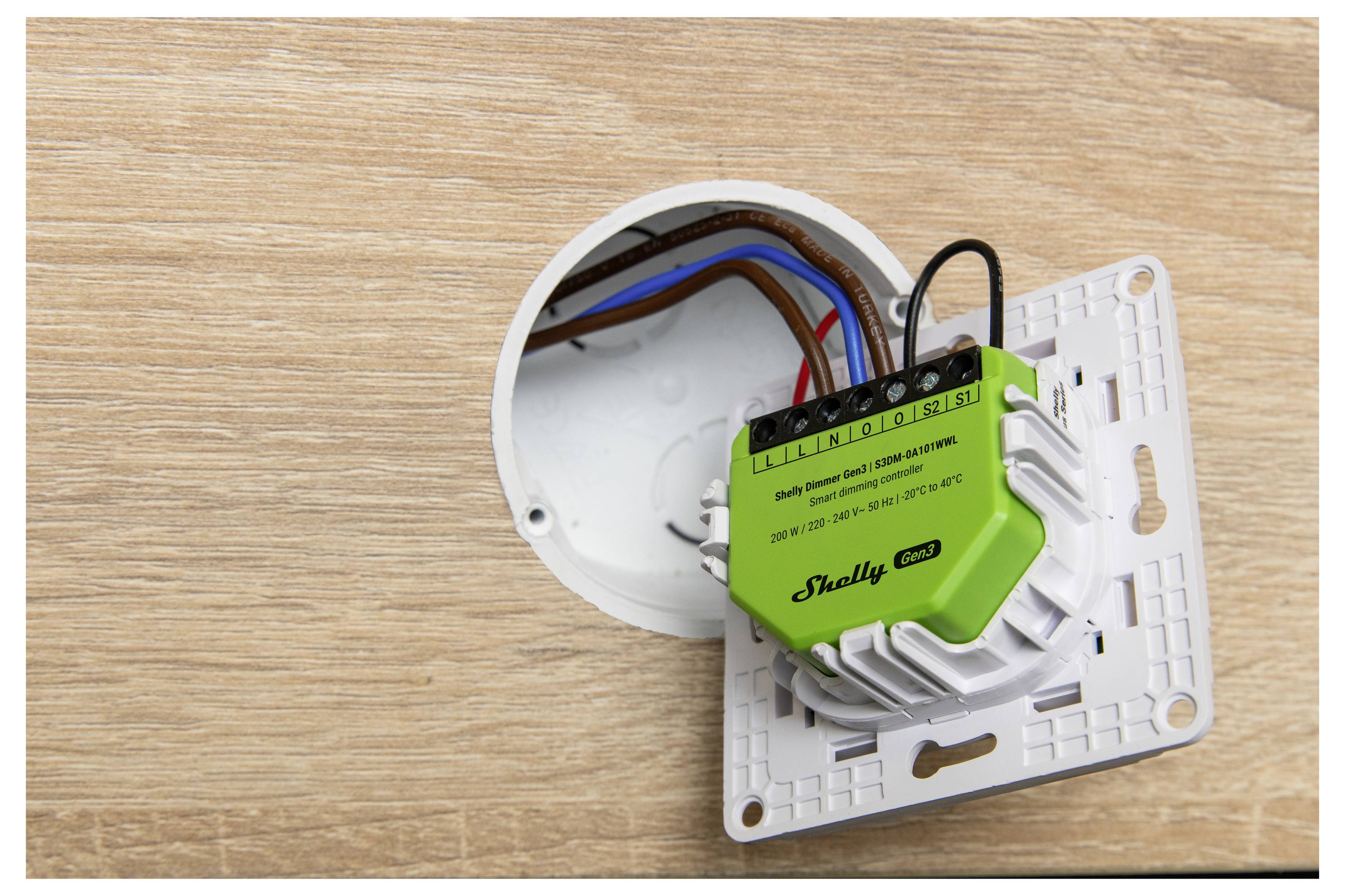 A green smart switch with wires is partially inserted into a wall socket against a wooden surface, demonstrating a home automation installation.
