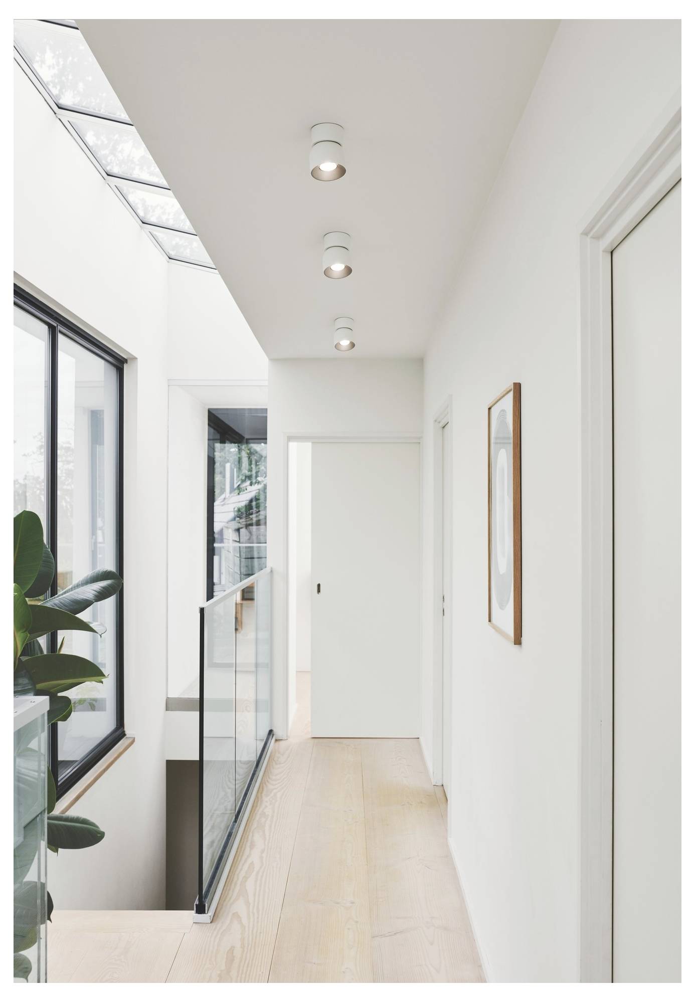 A modern, bright hallway with wooden flooring, glass railing, white walls, ceiling lights, and a large window on the left.