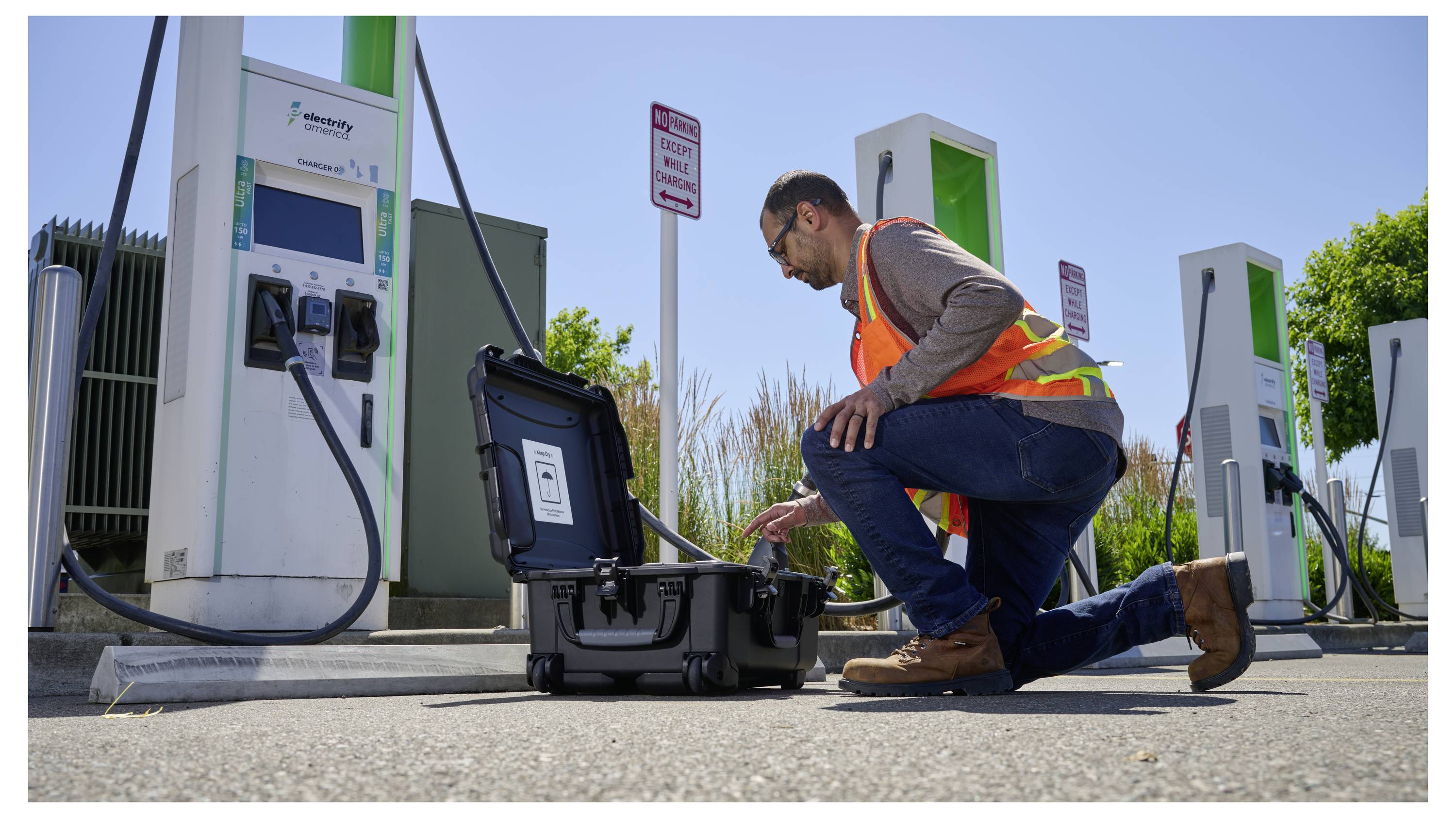 A technician in a safety vest kneels beside an electric vehicle charging station, examining a black equipment case, with two charging units in the background.