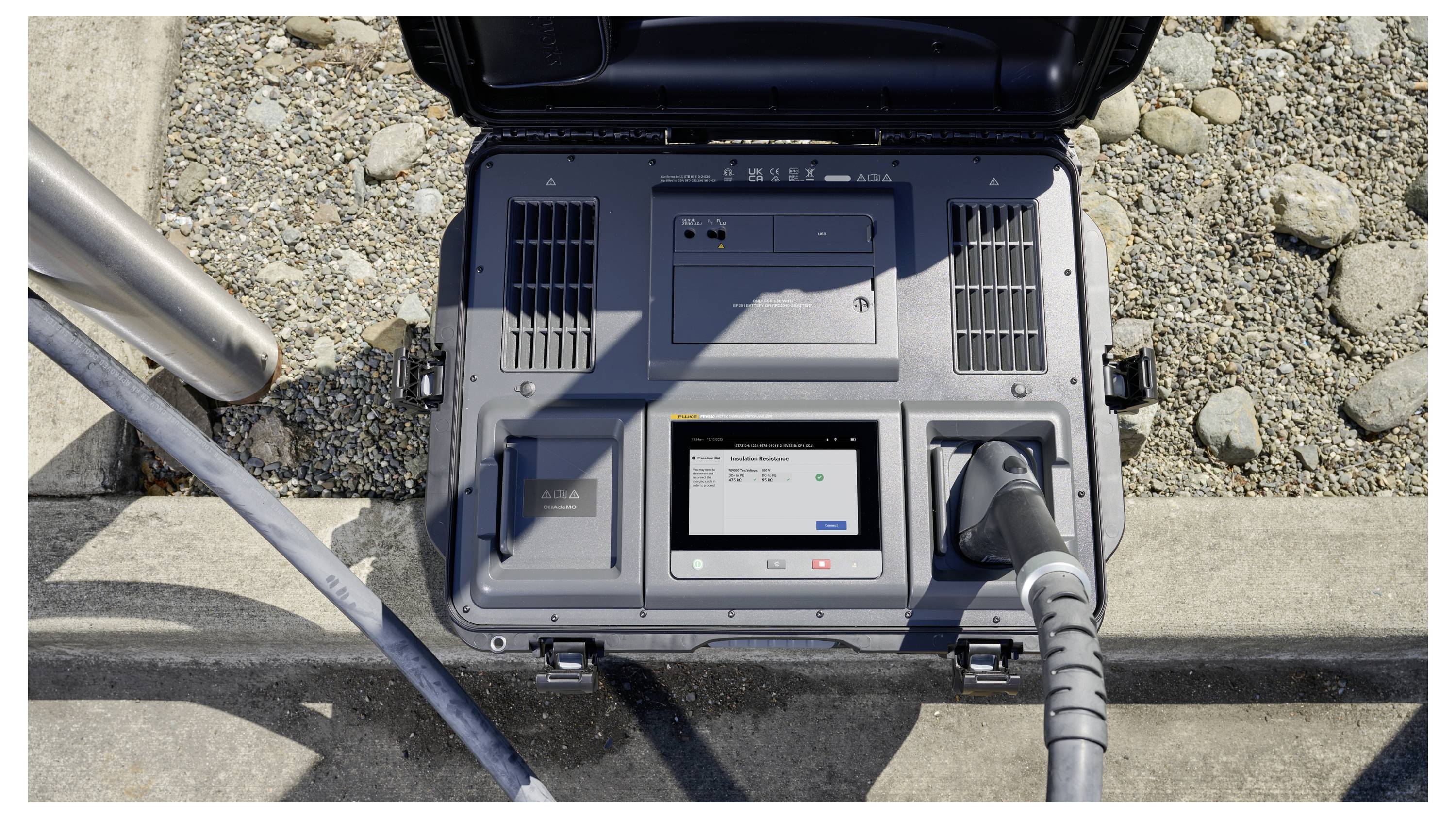 A portable electric charging station with a connected cable and digital display showing charging status, positioned on a rocky ground.