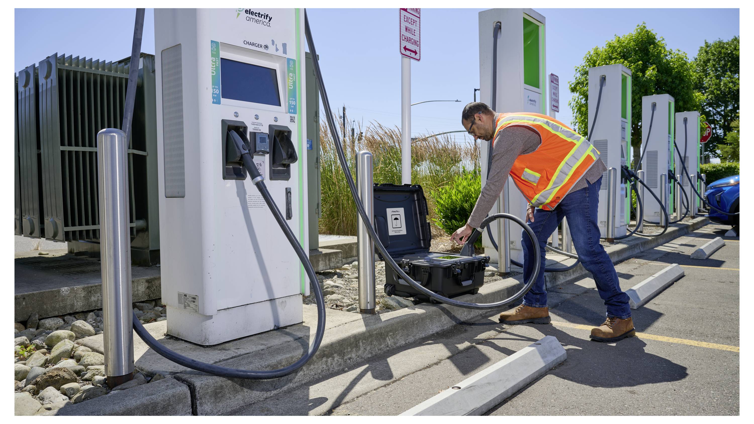 A worker in a safety vest connects a cable to an electric vehicle charging station outdoors, with cars and trees in the background.