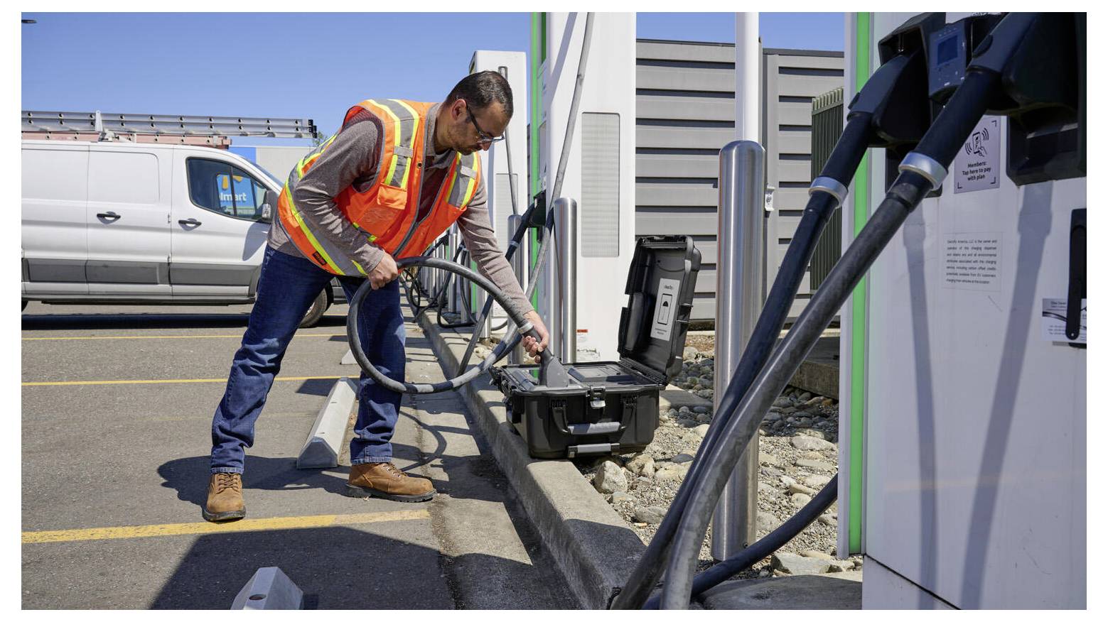 A worker in a safety vest plugs a charging cable into an electric vehicle at a charging station in a parking lot under clear skies.