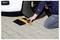 A person kneeling on a paved surface places a yellow measuring device near a car tire, likely checking pressure or alignment.