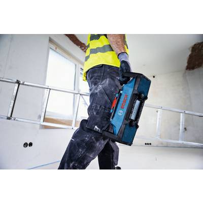 A worker in construction attire holds a Bosch tool case, walking through an unfinished room with a ladder and patches on the walls.