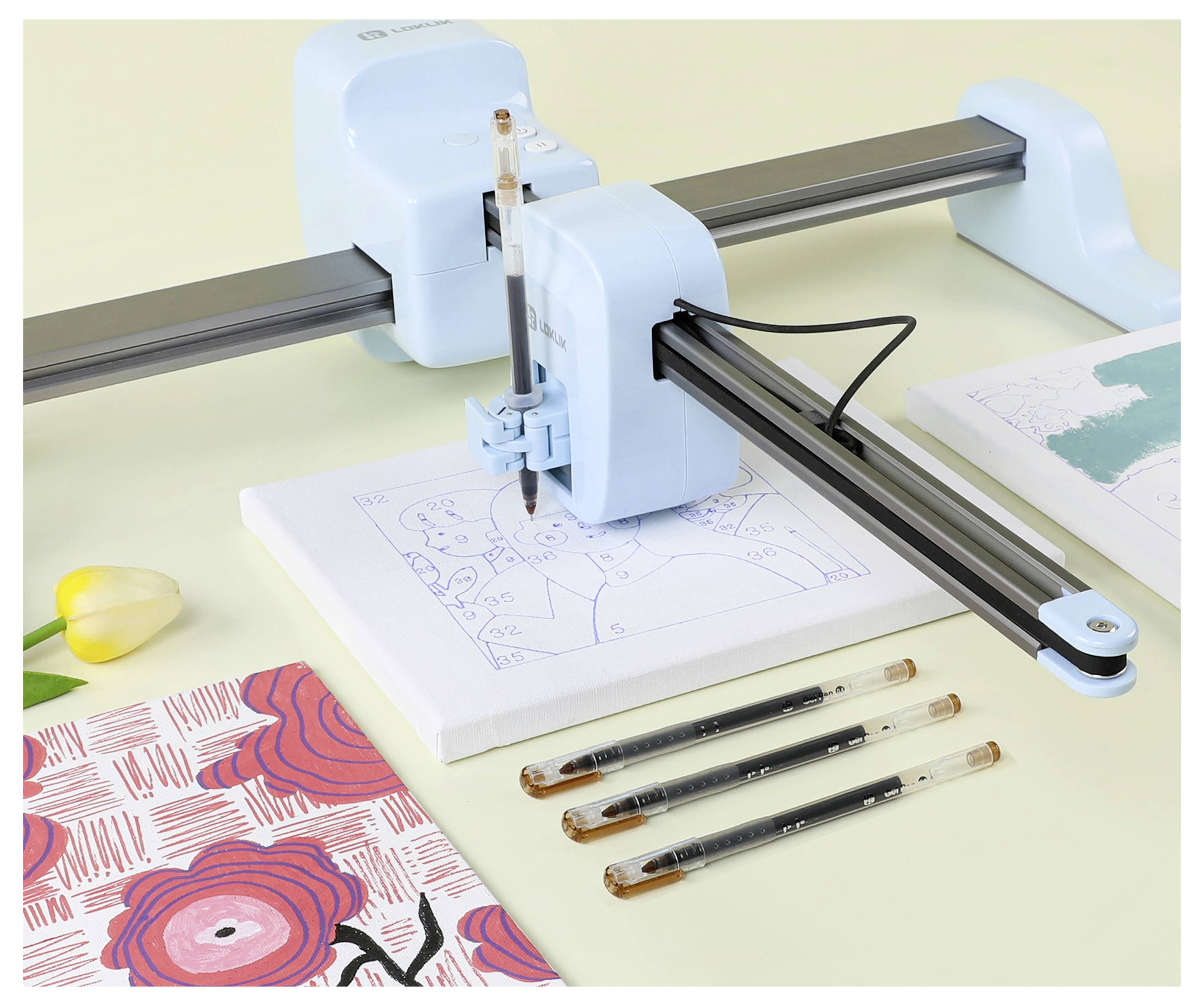 'A drawing machine sketching an illustration of two people on canvas with pens and a floral pattern book nearby on a table.'
