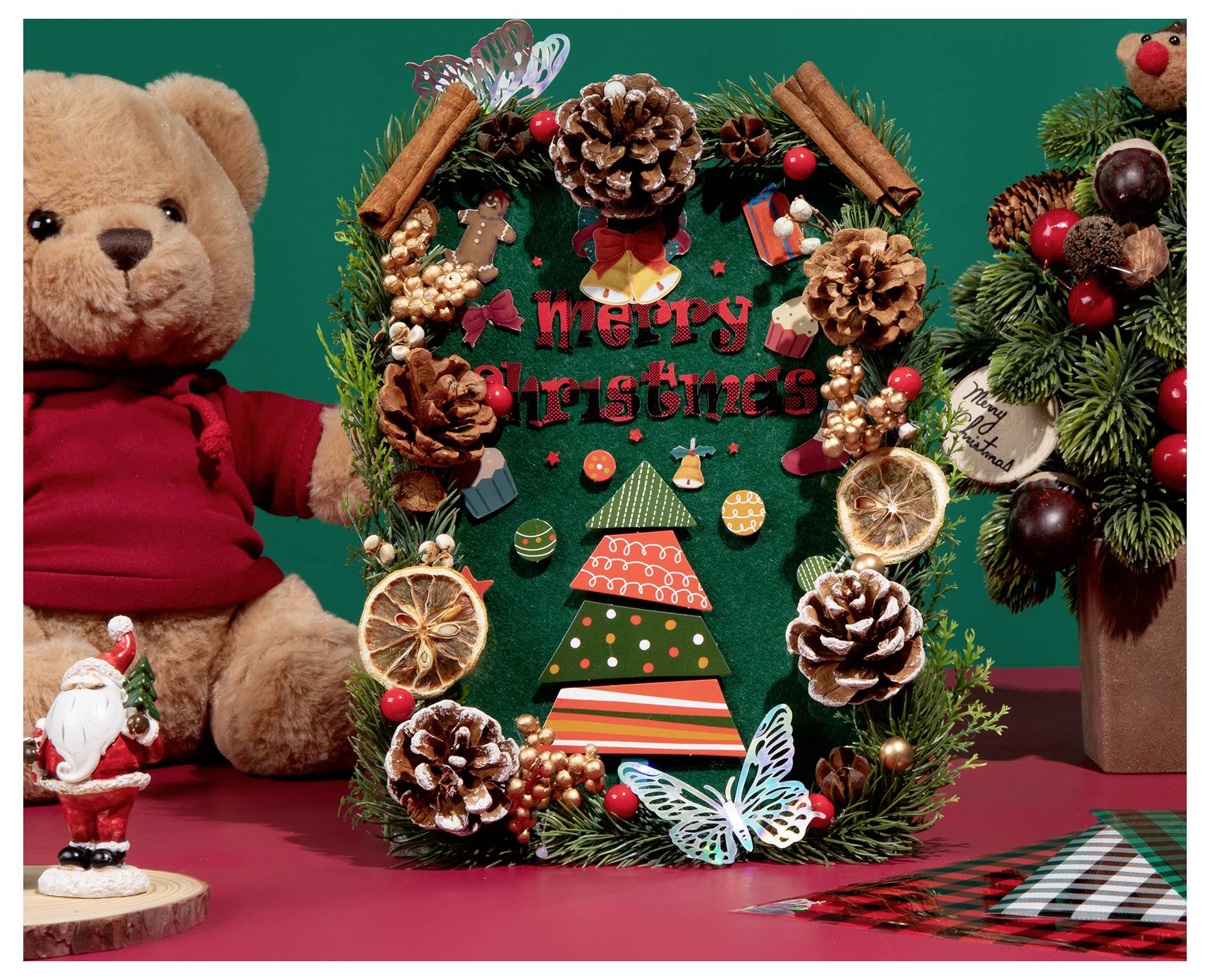 Festive holiday display with a 'Merry Christmas' sign surrounded by pinecones, dried fruit, and decorations. Teddy bear and Santa figure in view.