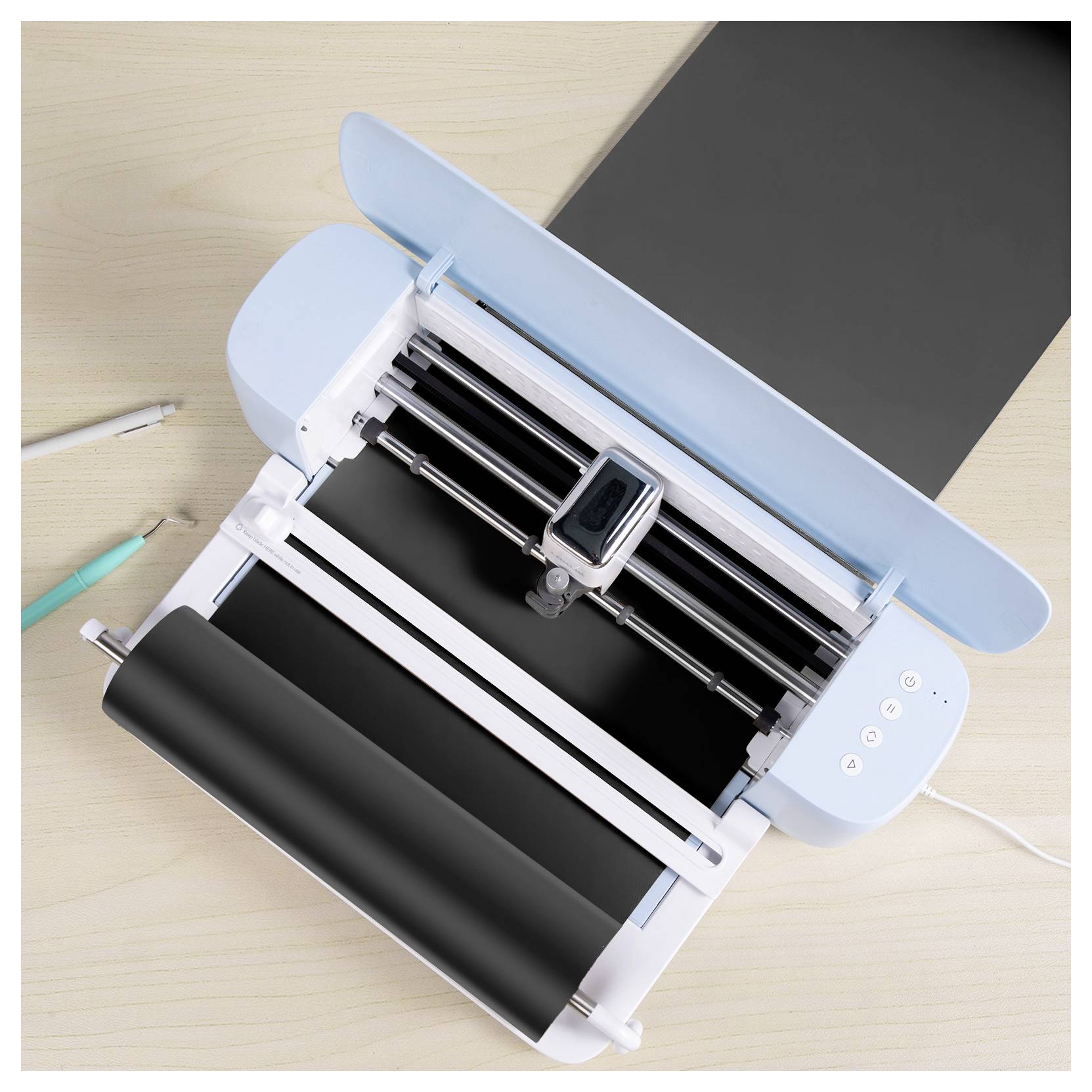 A cutting machine with a blue lid is actively cutting black vinyl on a light wood table, with a pen and marker nearby.