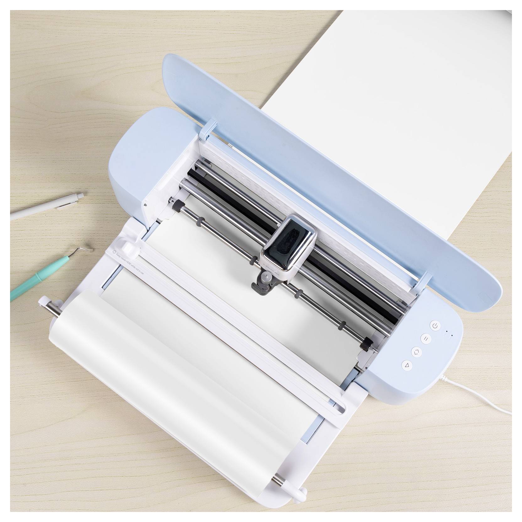A tabletop cutting machine with a light blue lid, holding white paper. Two pens are placed nearby on a wooden surface.