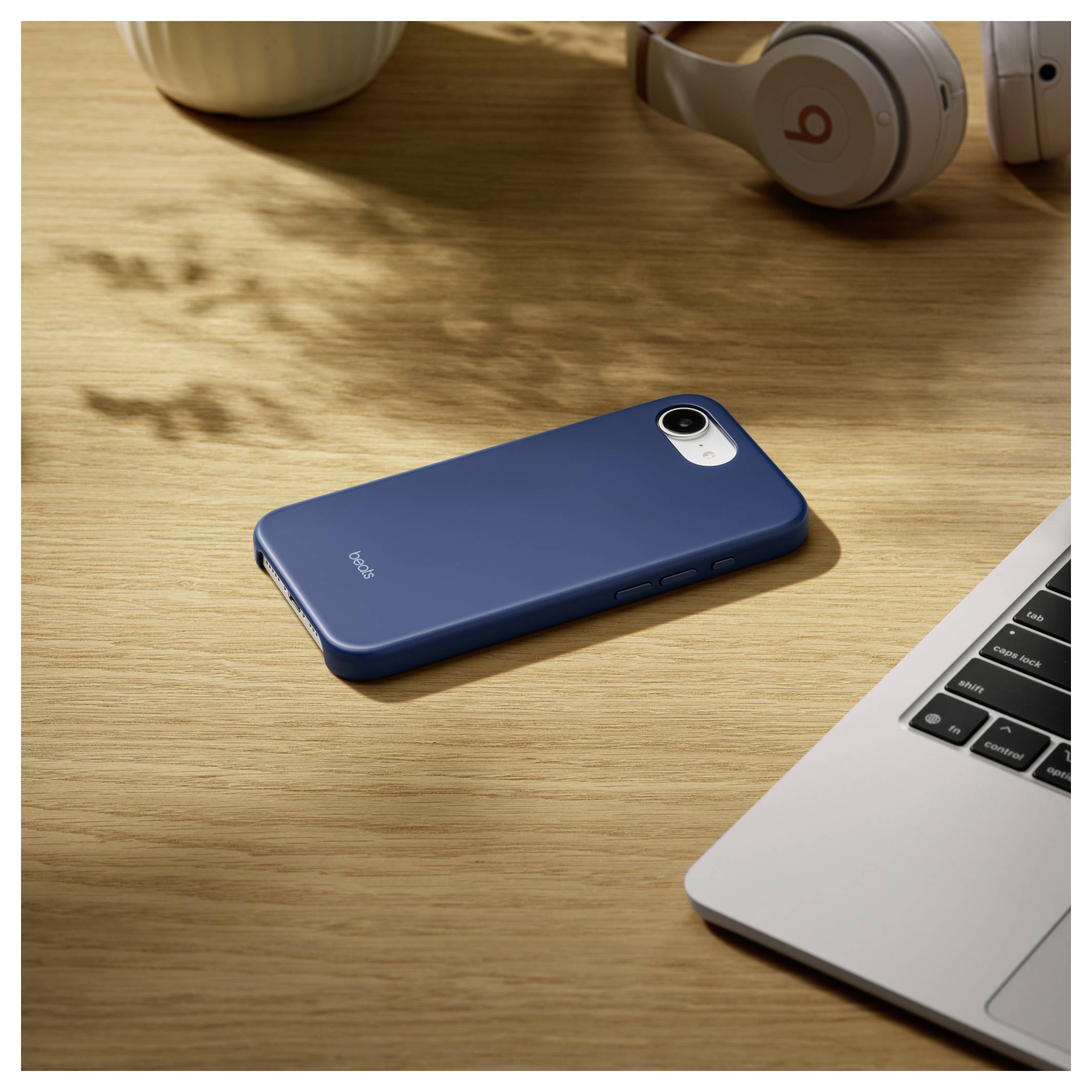 A blue smartphone with a protective case lies on a wooden desk next to a laptop and headphones.