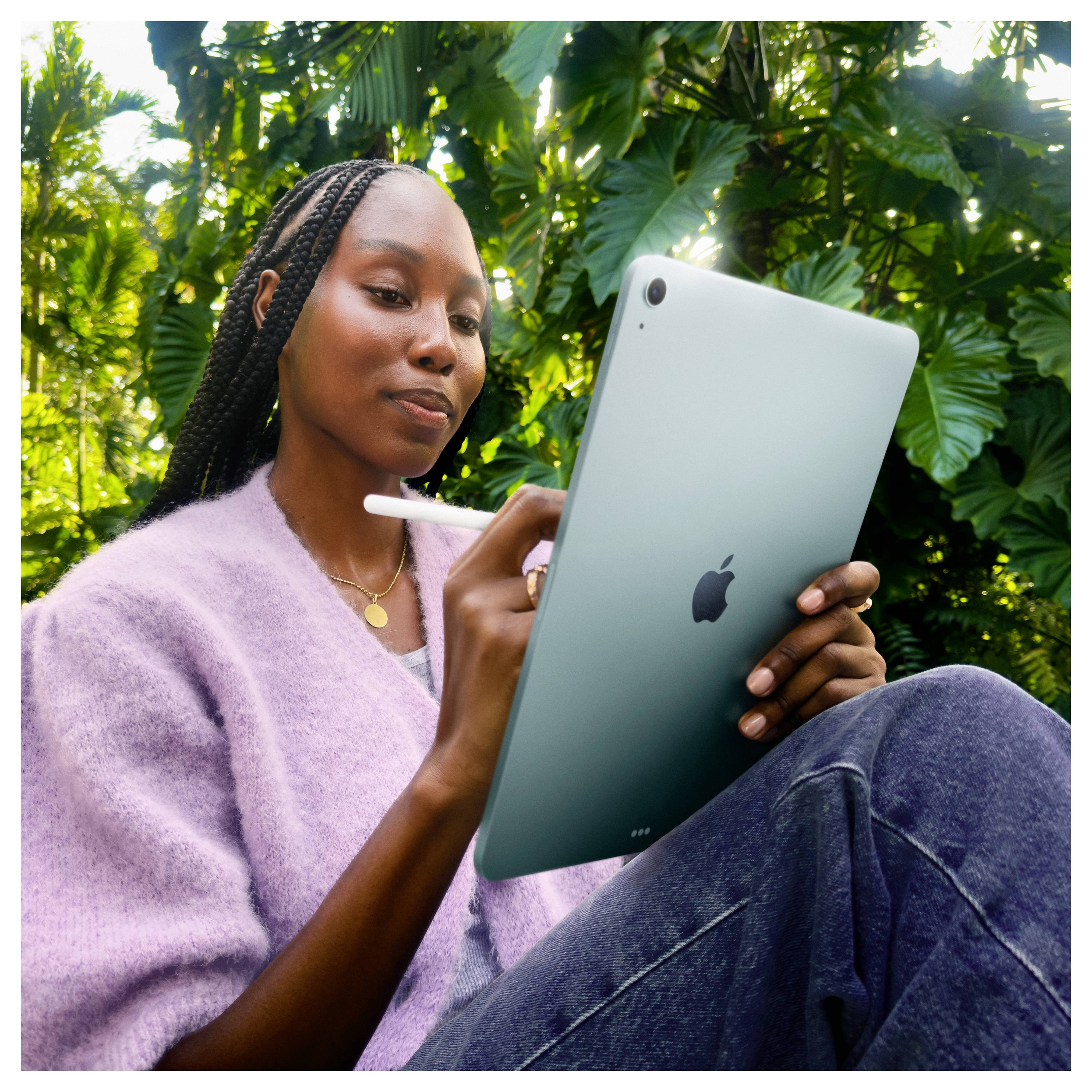 A person using a tablet with a stylus, sitting outdoors in a garden setting, surrounded by lush green foliage, focused on the screen.