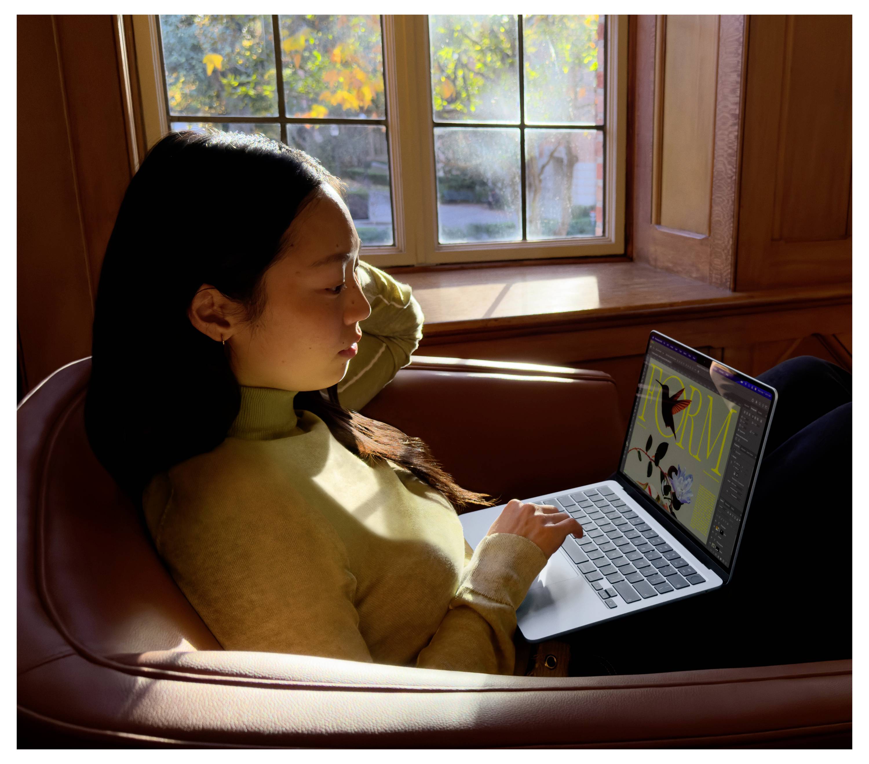A person is sitting in a chair in front of a window and working on a laptop. Autumn trees are visible outside.