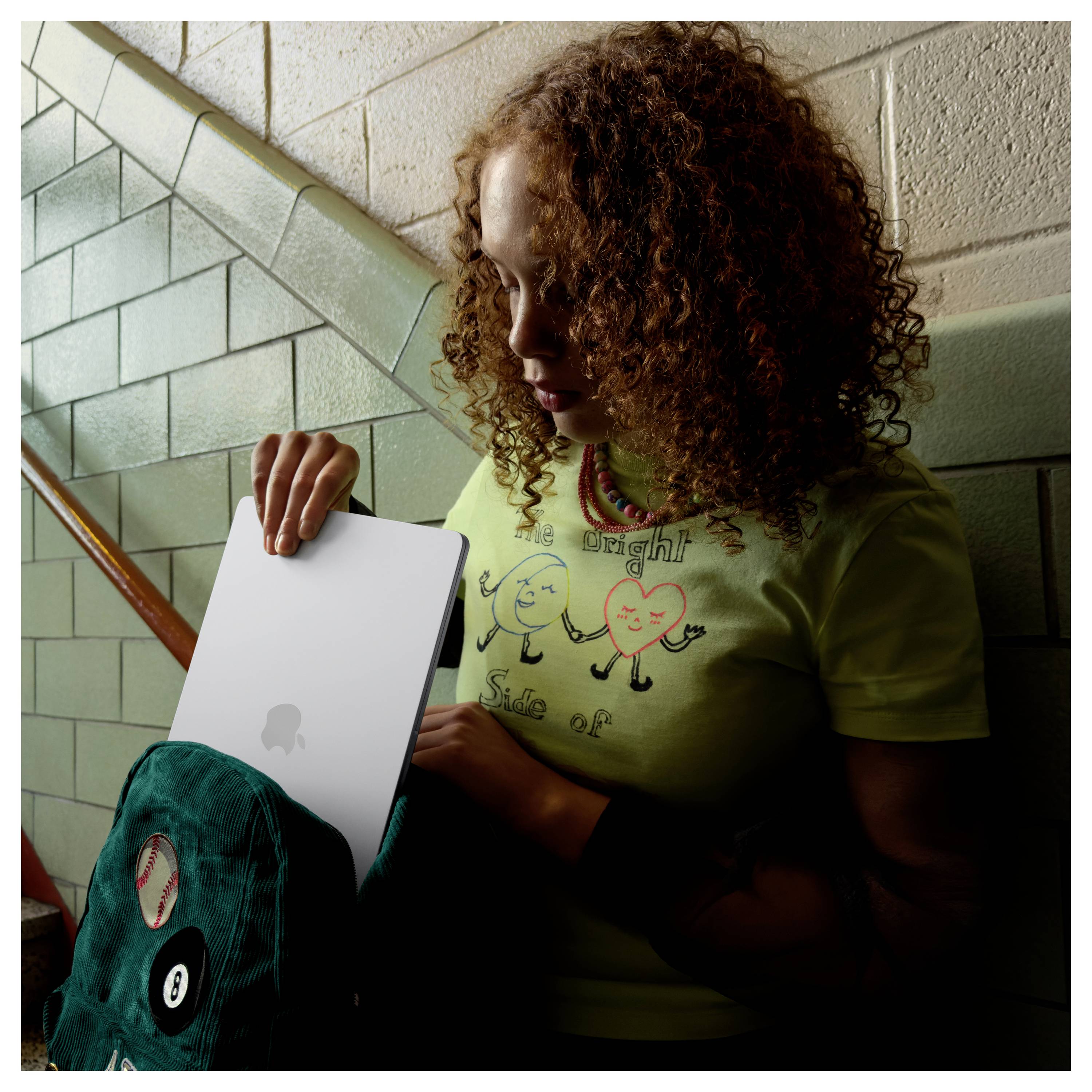 A person with curly hair packs a laptop into a backpack. The person wears a green shirt with cartoon characters. They're next to a tiled wall.