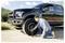 Man kneels beside a black pickup truck, using a hand pump to inflate a front tire on a roadside.