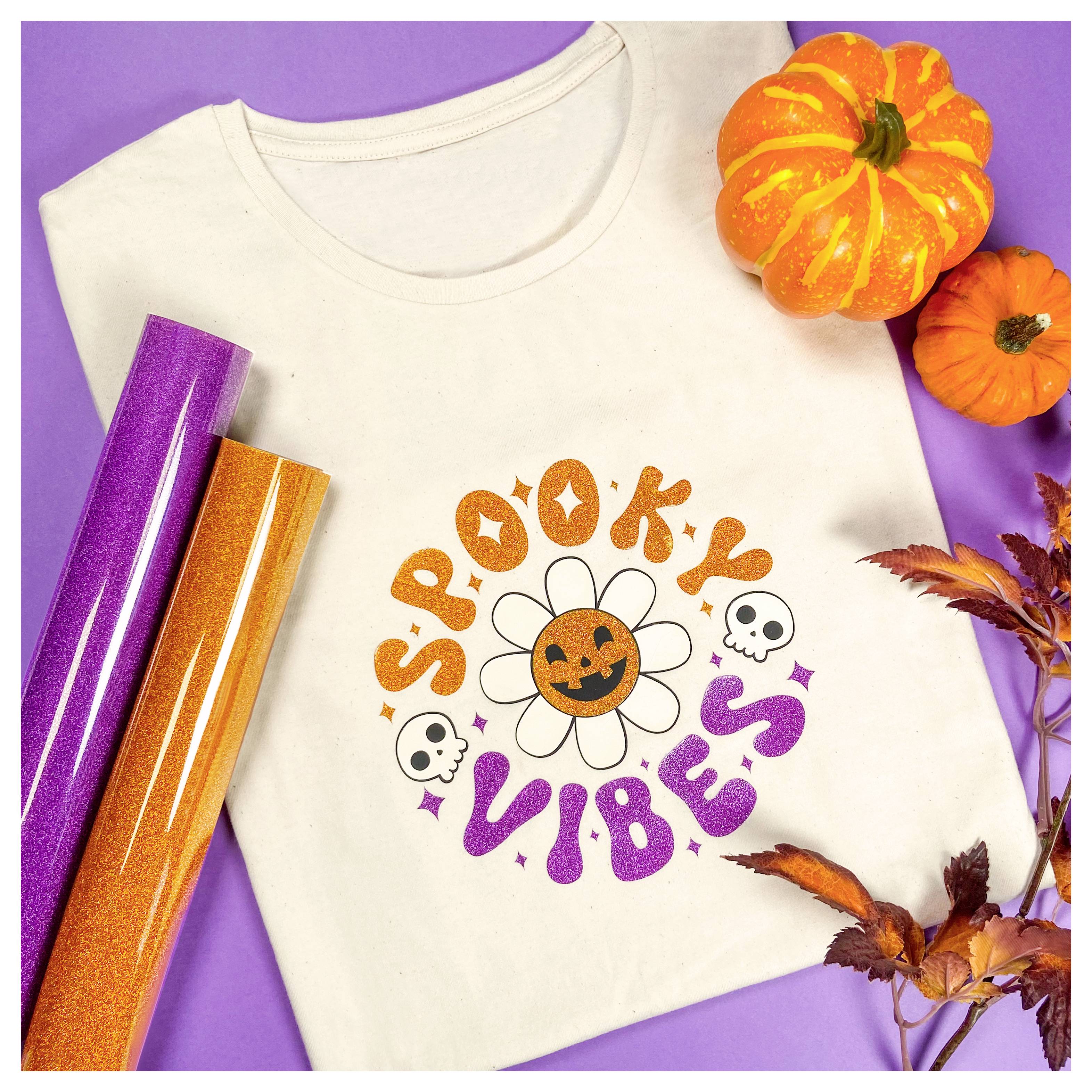 A white t-shirt with the text 'Spooky Vibes' featuring a smiling pumpkin and two skulls, surrounded by orange and purple Halloween decorations.