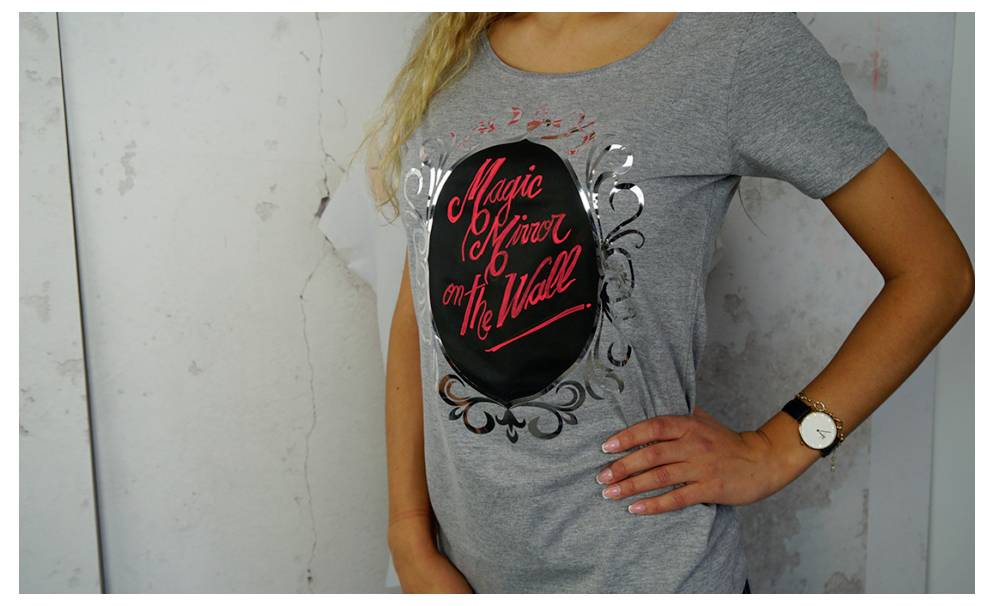 A person wearing a gray t-shirt with a red and black design reading 'Magic Mirror on The Wall,' standing against a light-colored wall.