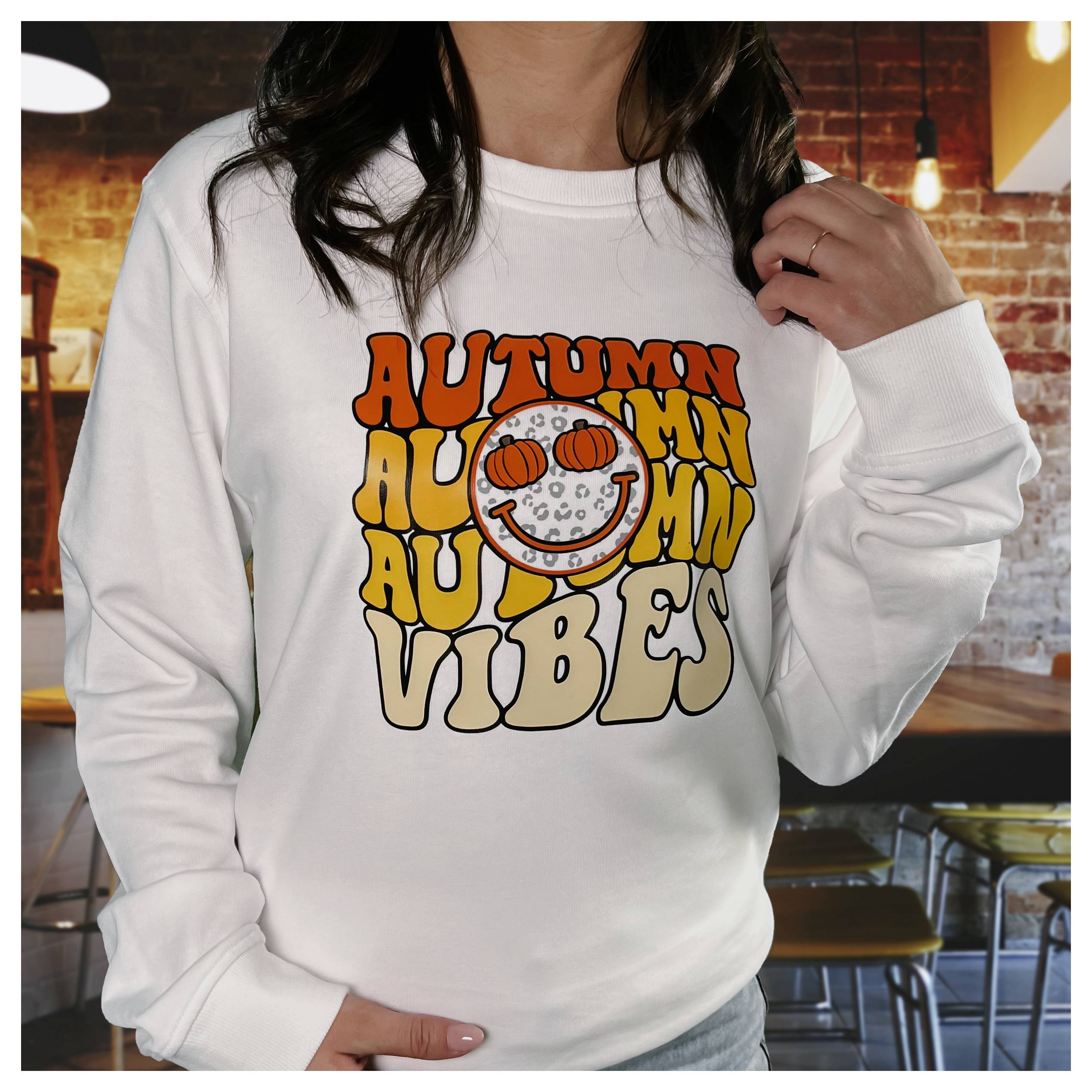 A person wearing a white sweatshirt with a colorful 'Autumn Vibes' design featuring a smiling pumpkin face, standing in a cozy indoor setting.
