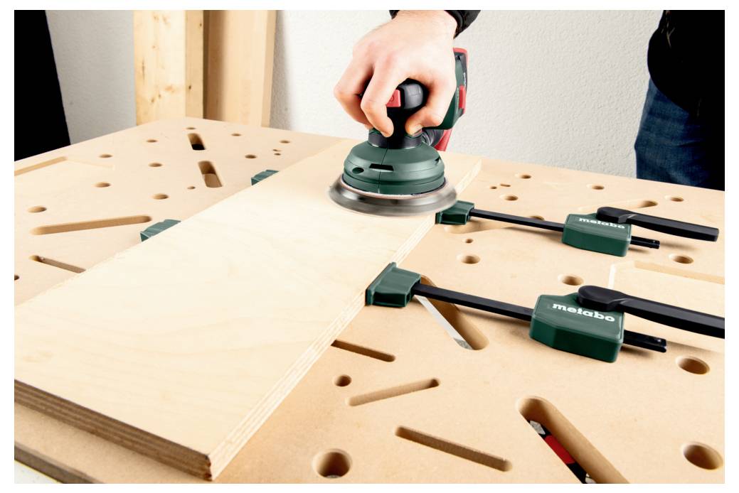 A person sands a wooden plank with an orbital sander on a workbench, secured by green clamps, emphasizing woodworking or DIY activity.