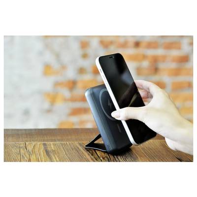 A hand holds a smartphone over a wireless charging stand on a wooden surface, with a brick wall in the background.