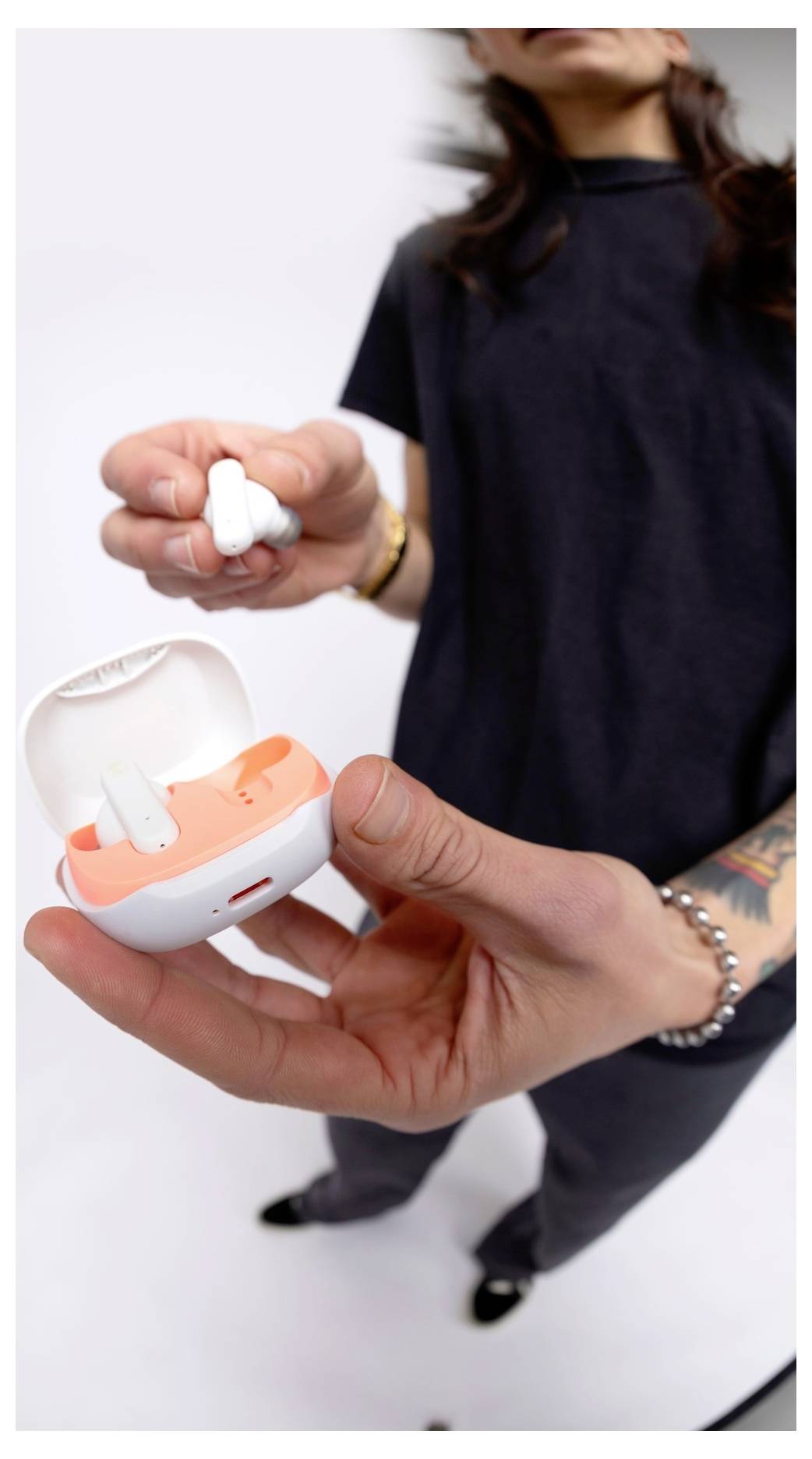 A person holds an open white and orange earbud case with a single earbud in hand, wearing casual dark clothing on a light background.