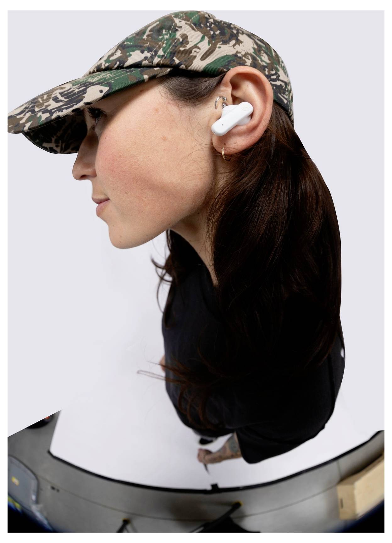 A person wears a camouflage cap and white wireless earbuds, looking to the left. The image is distorted, emphasizing facial features and cap.