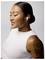 A person with earbuds smiles, wearing a sleeveless white top. Their arm has a tattoo, and hair is styled in a neat bun.