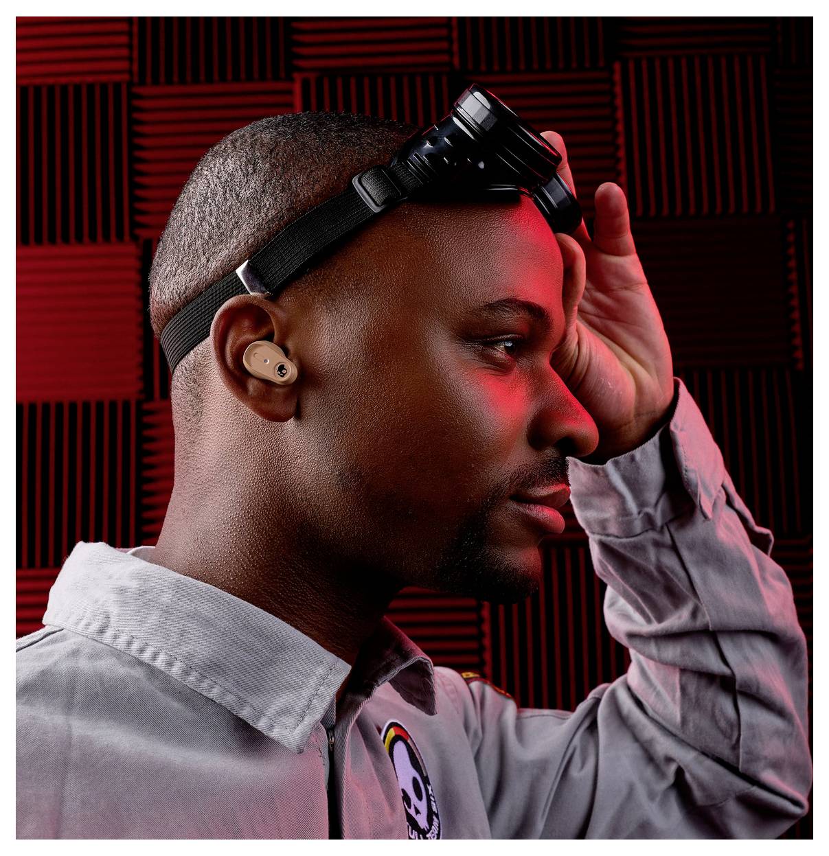 A person wearing headphones and a headset with goggles in a red-lit room lined with acoustic foam, looking contemplative to the side.