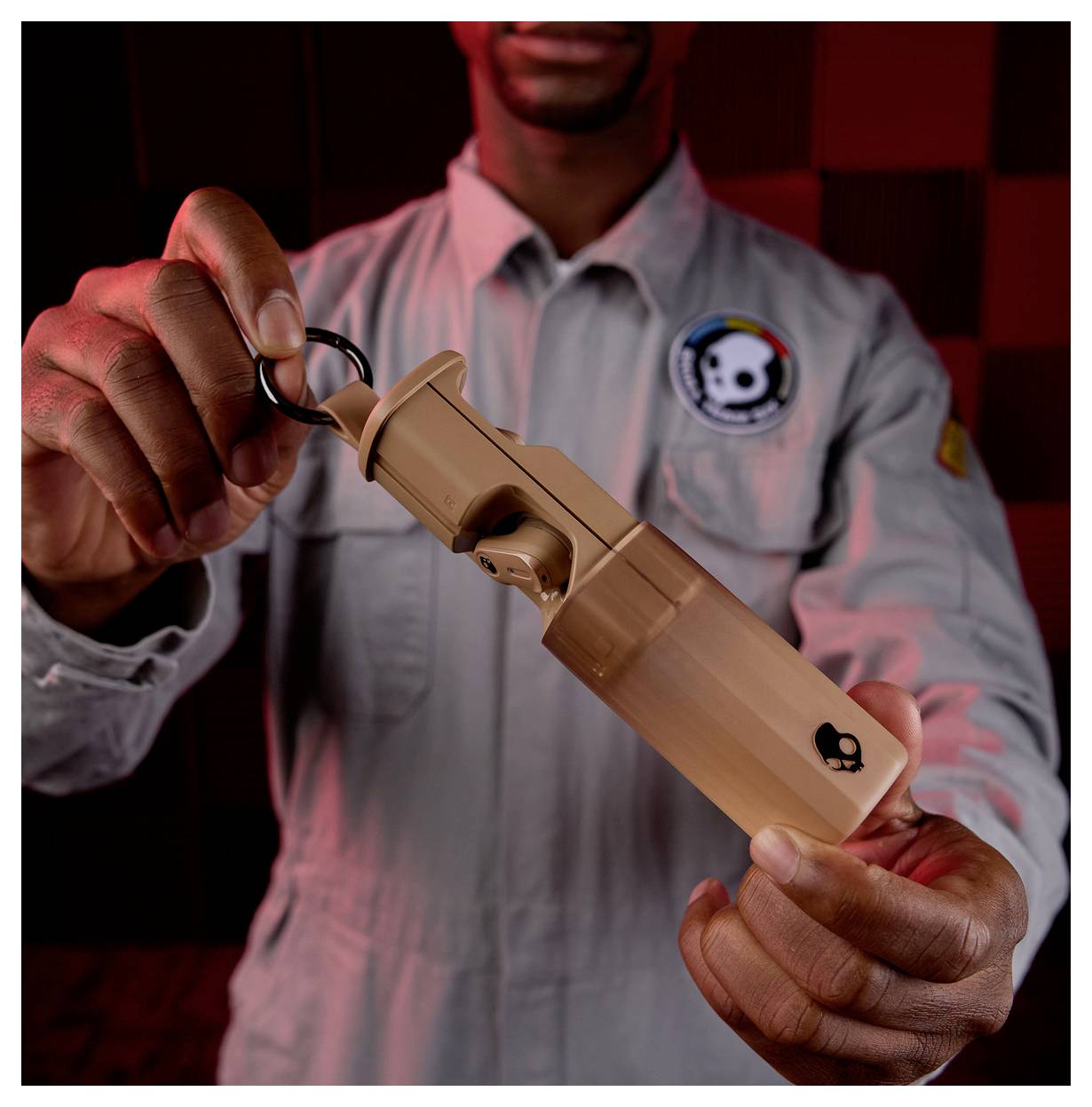 A person in a uniform holds a tan bottle-like object with a skull logo, under red lighting.