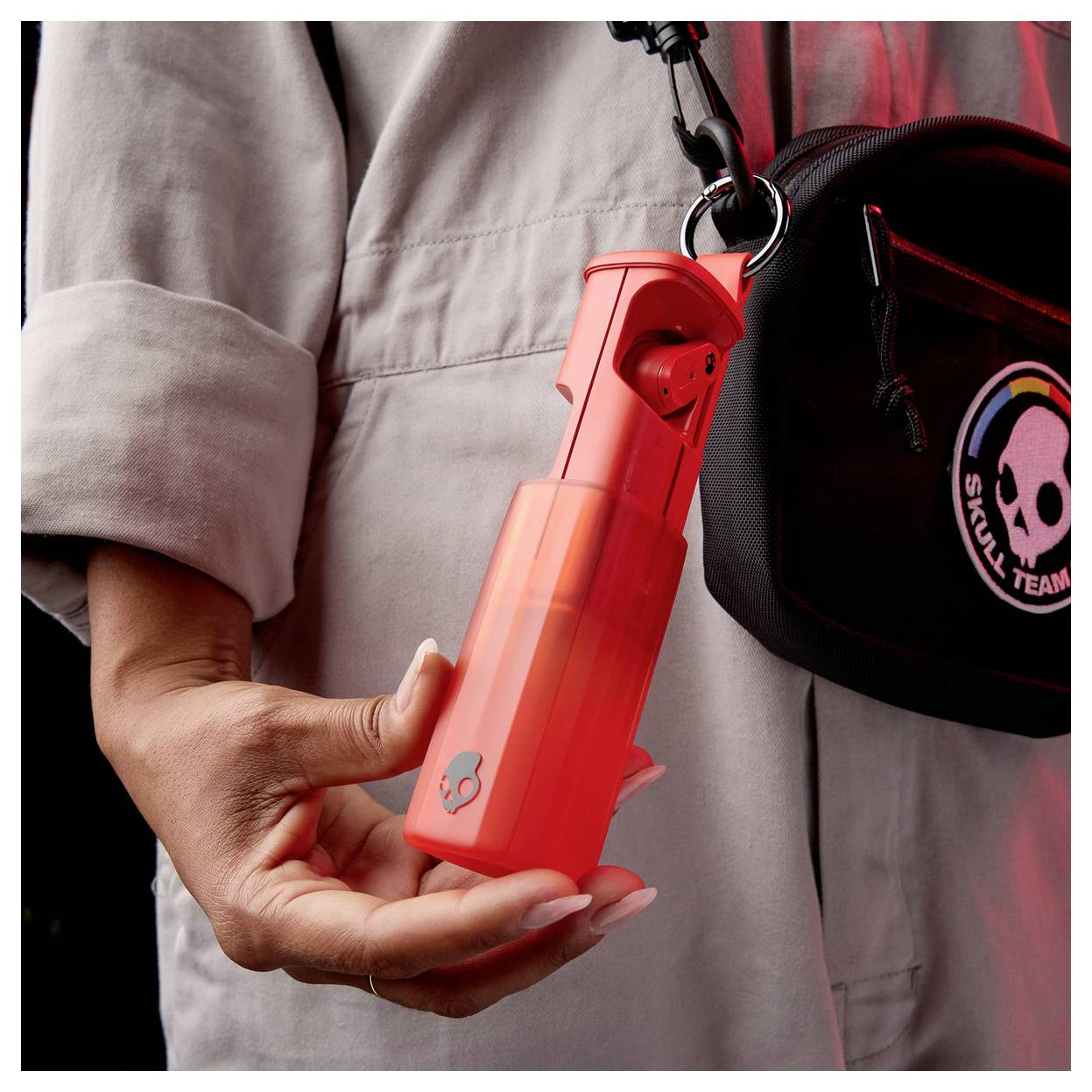 A person holds a bright red pepper spray canister attached to a black bag with a 'SKULL TEAM' patch, suggesting readiness for safety.
