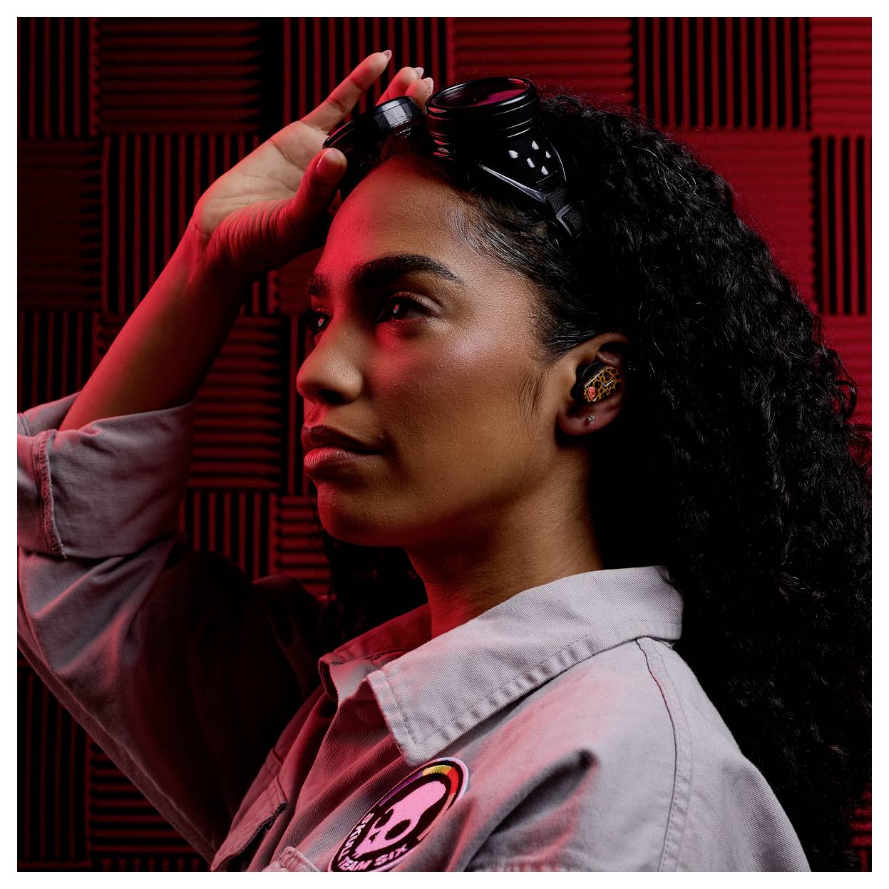 A person wearing protective goggles and a grey jacket in a soundproof room, lit with red light, looks to the side in a contemplative pose.