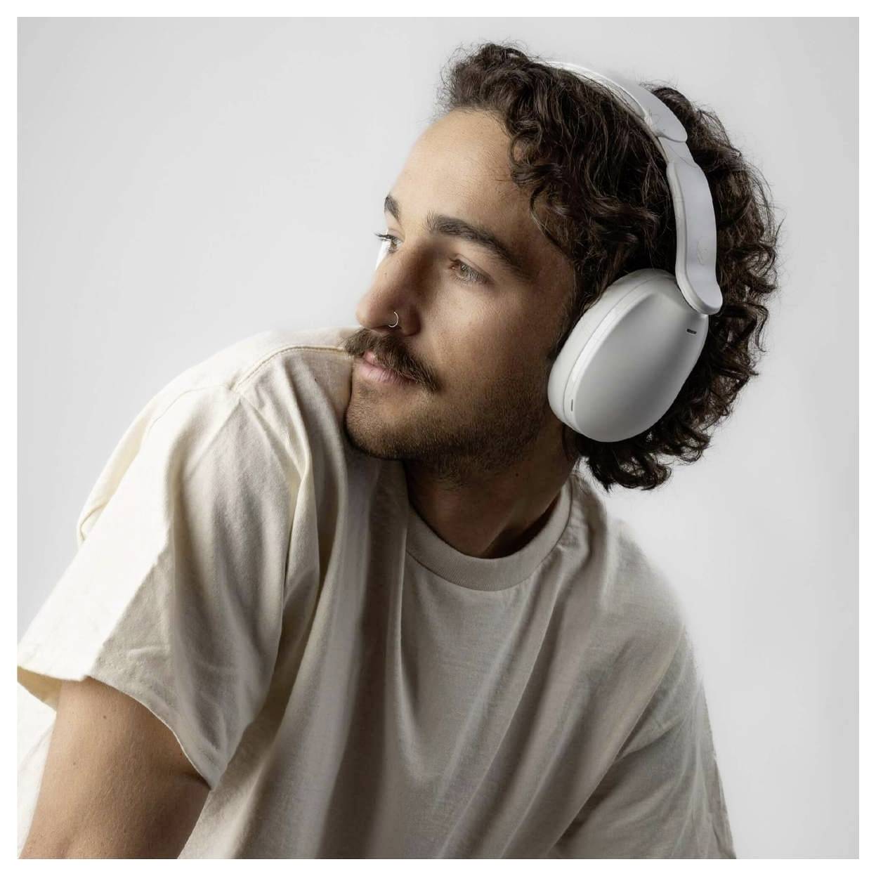 A person wearing white headphones and a beige t-shirt looks to the side against a plain background.